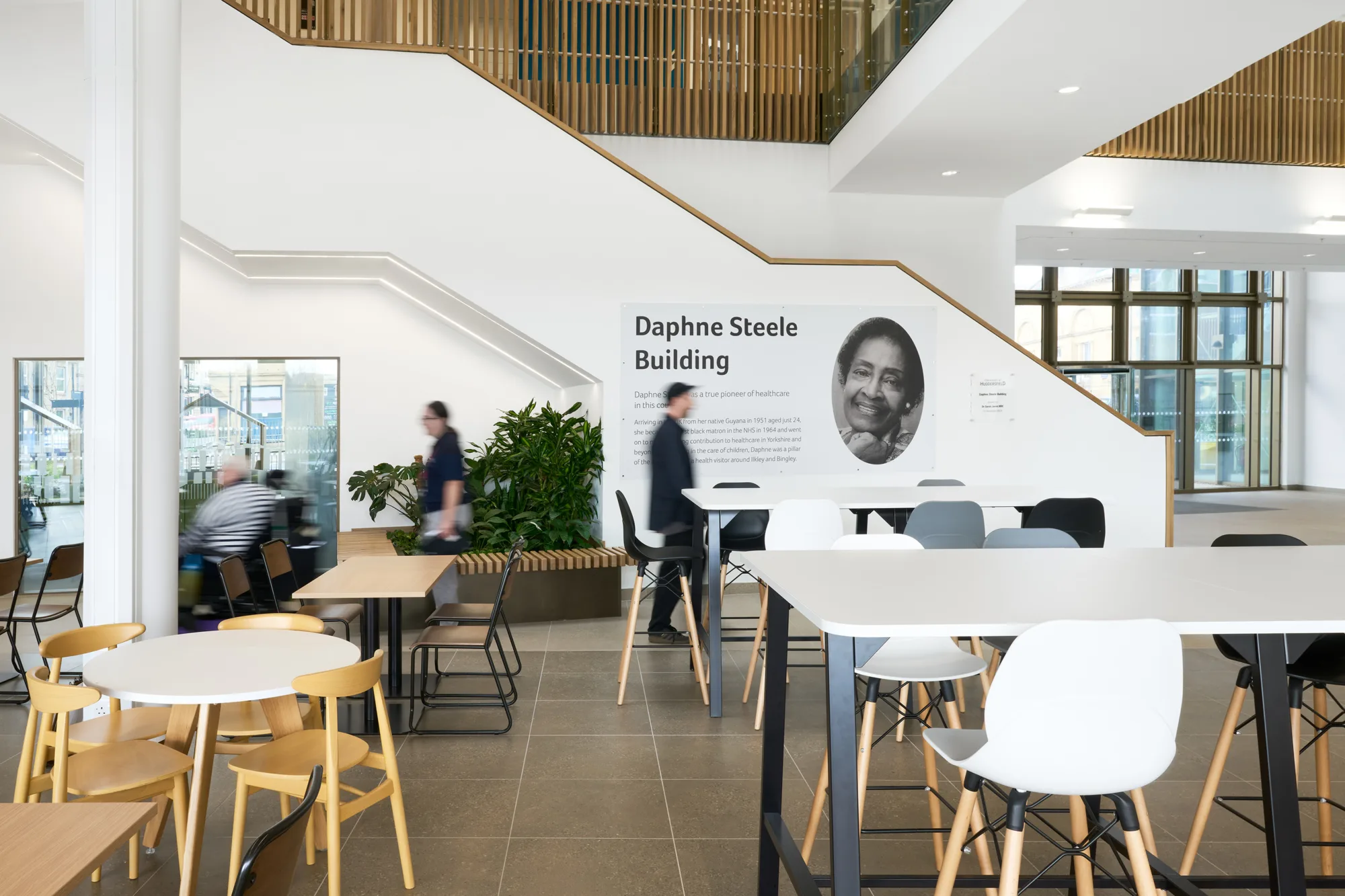 A modern indoor space with tables and chairs, a staircase to an upper level, and a wall sign reading "Daphne Steele Building" beside an oval mirror; people are walking and sitting.