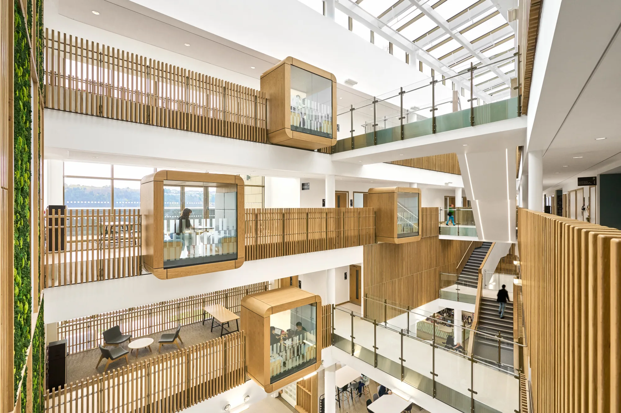 A multi-story atrium with wooden accents, glass railings, suspended wooden cubicles with glass walls, open seating areas, staircases, and a vertical green garden on the left wall.