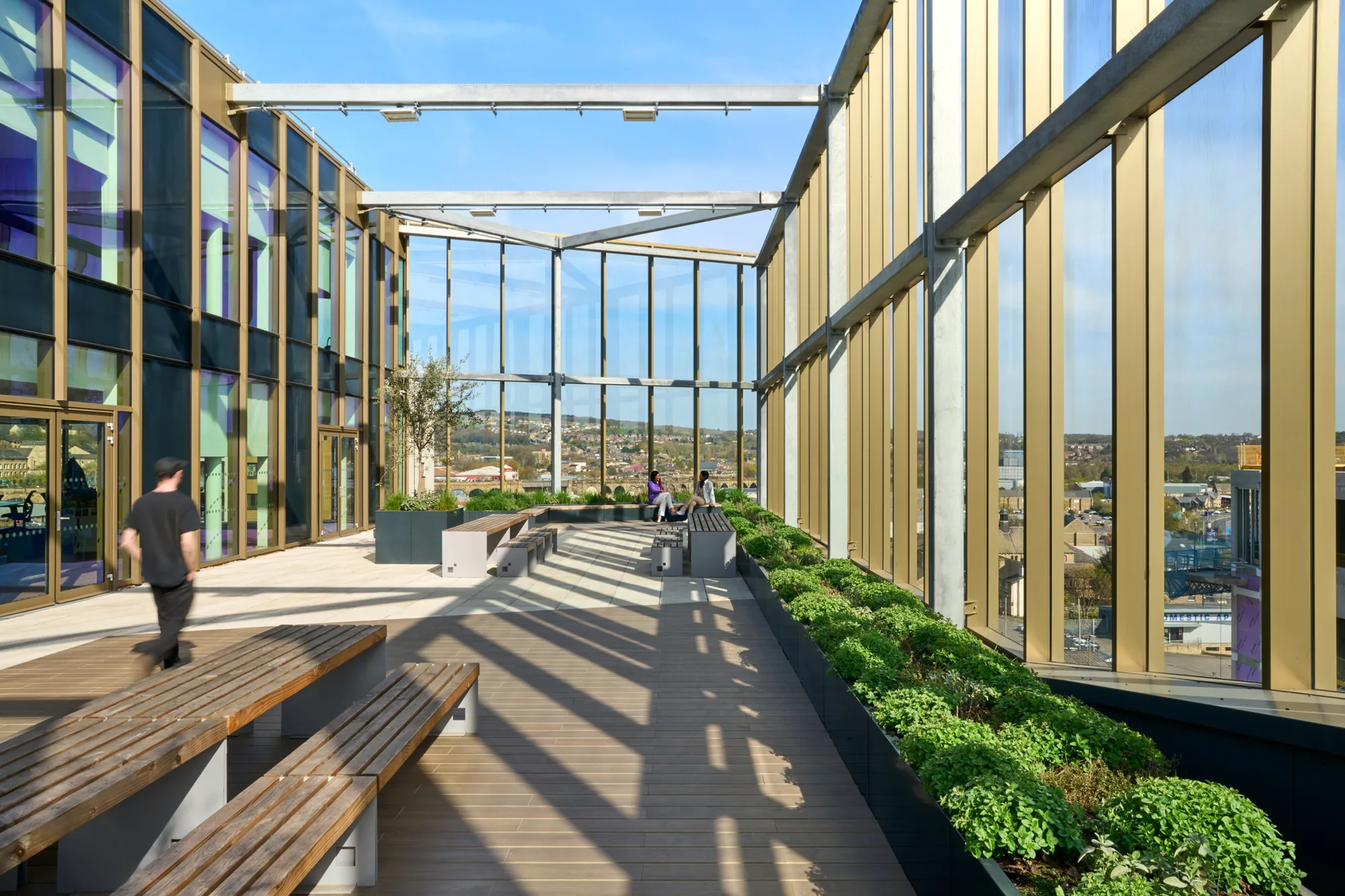 A rooftop terrace with wooden benches, planters containing green shrubs, and a few people in the background; enclosed by glass walls and metal beams with a cityscape visible beyond.