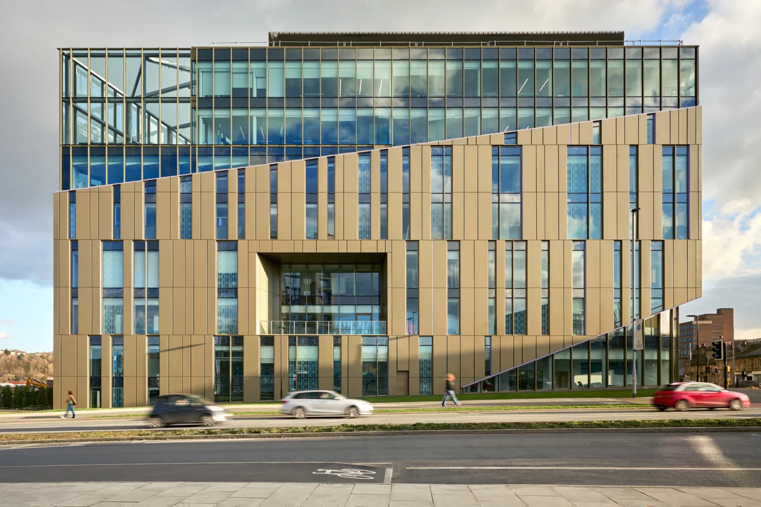 A multi-story building with vertical beige and blue panels, large glass windows, and an angular facade; cars drive on the road and pedestrians walk on the adjacent pavement.