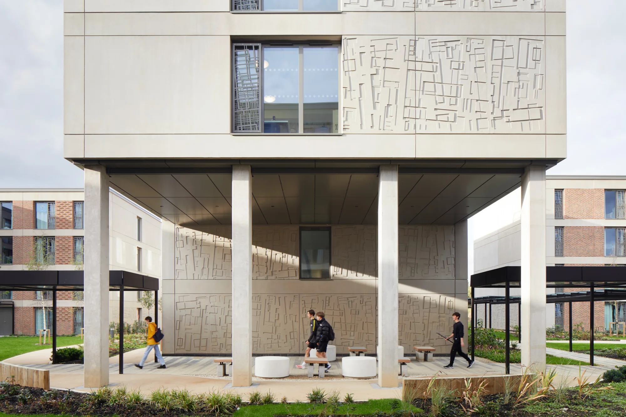 A modern building with patterned exterior panels stands on columns above a sheltered walkway. Three people walk beneath the structure near round seating and planted landscaping, with additional multi‑story buildings visible in the background.