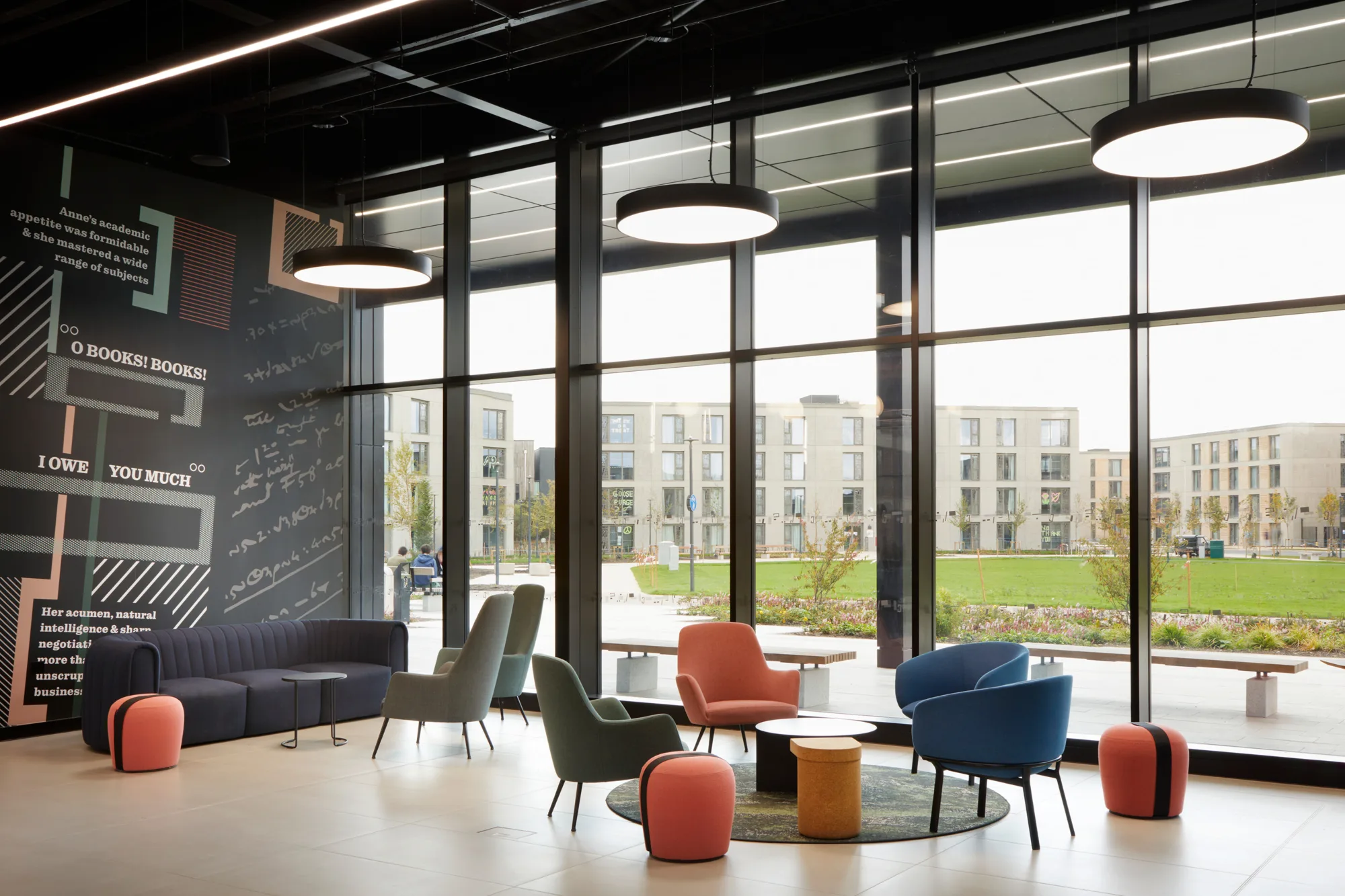 A seating area with sofas, chairs, small tables, and round ceiling lights is shown beside a floor‑to‑ceiling window. A wall with graphic patterns and text is on the left, and modern multi‑story buildings and lawns are visible outside.