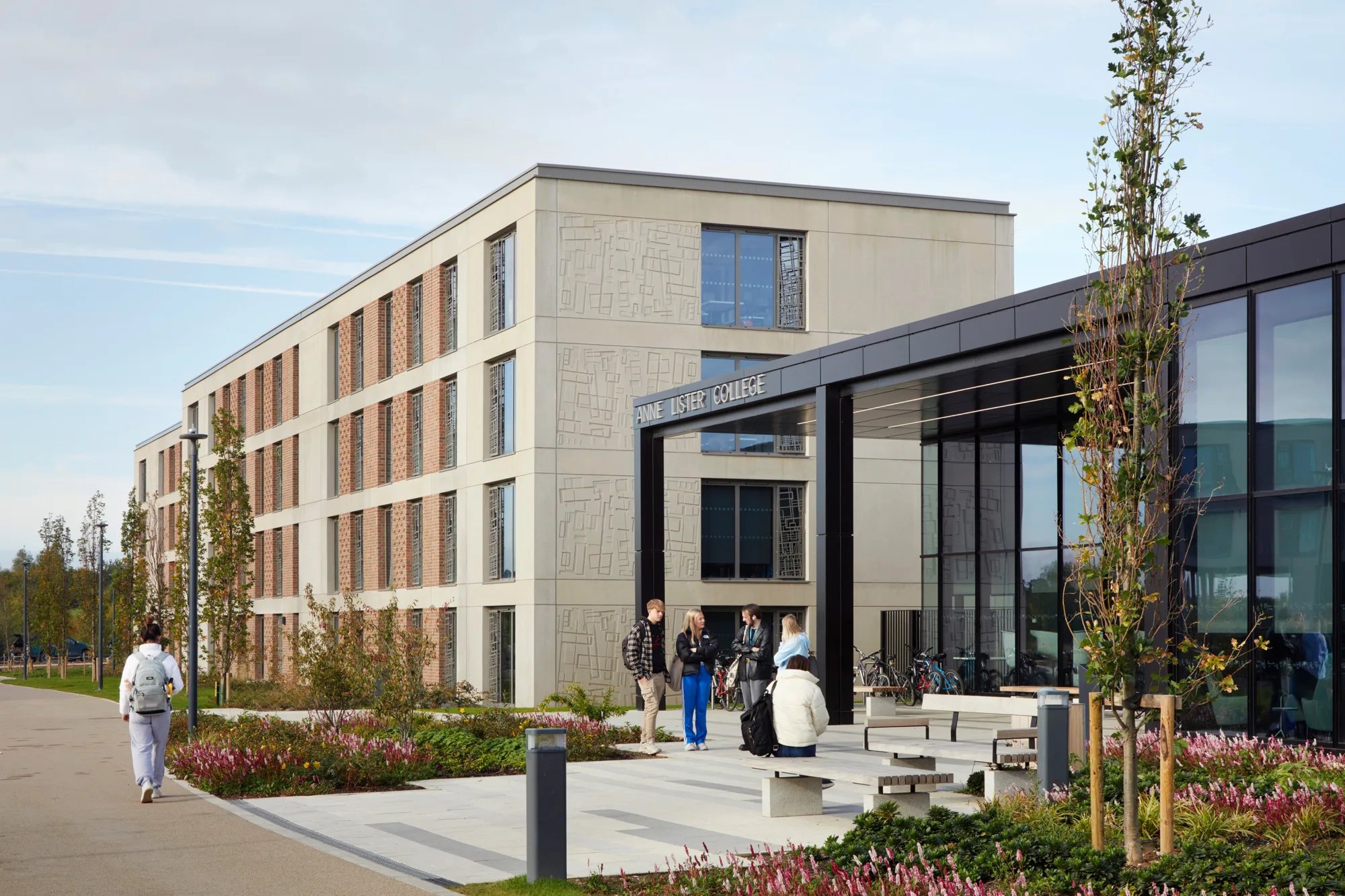 A modern multi‑story building with brick and light‑coloured panels stands beside a glass‑fronted entrance marked “ANNE LISTER COLLEGE.” Several people walk or stand near benches, landscaping, and a paved pathway in the foreground.