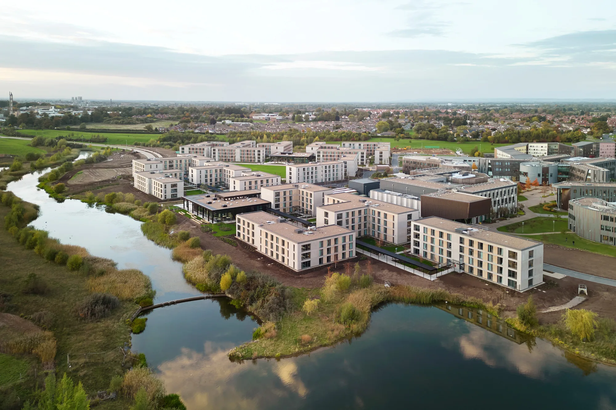 Aerial view of modern multi‑story buildings arranged around pathways and green areas, next to a reflective body of water with surrounding vegetation. Additional buildings and residential areas extend into the distance.