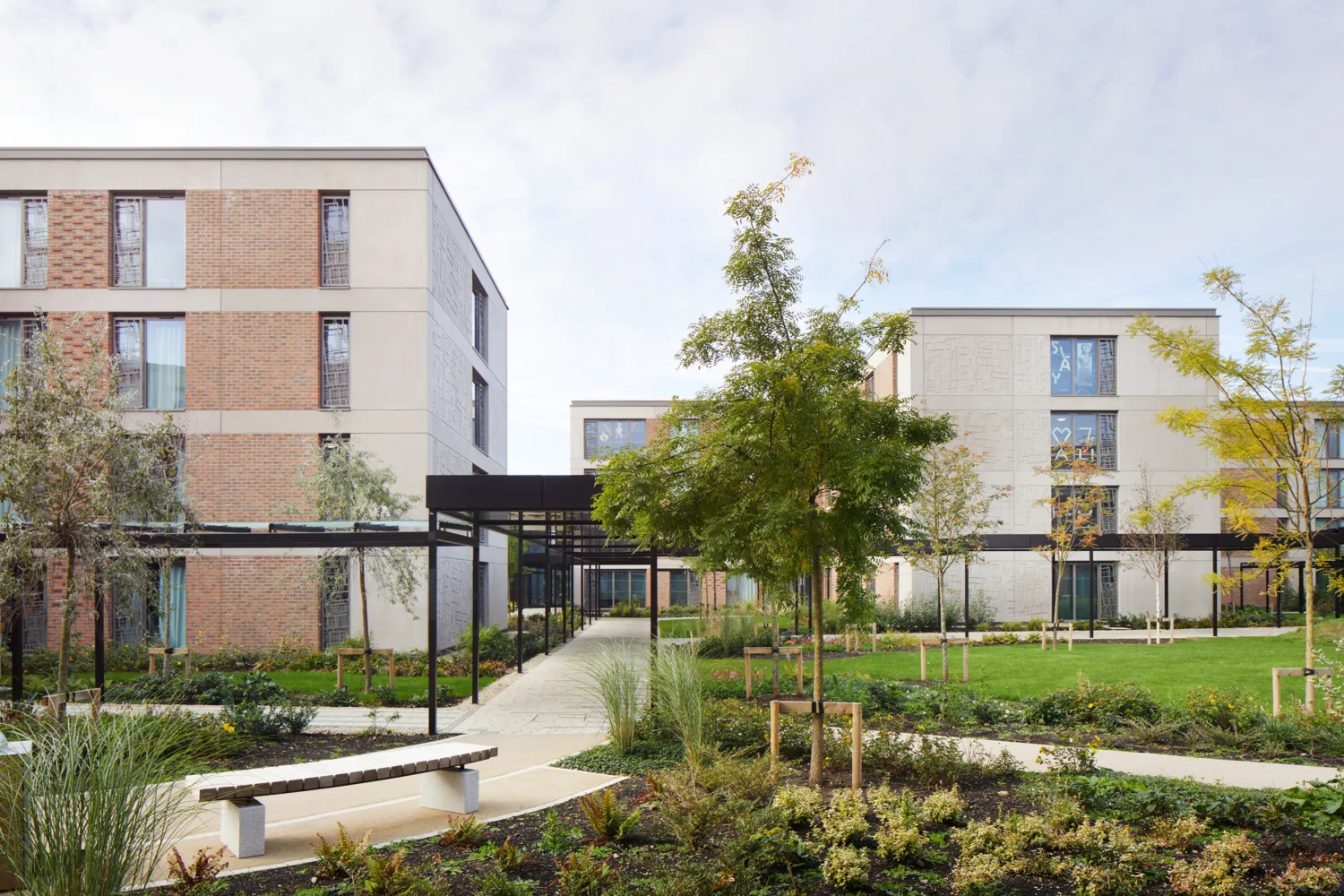 A landscaped courtyard with paved walkways, benches, and planted trees surrounded by modern multi‑story buildings with brick and light‑coloured facades, large windows, and metal walkway structures.