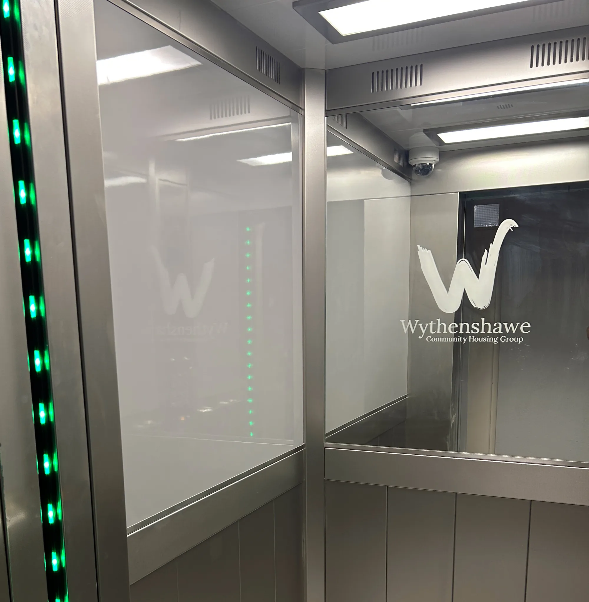Interior of a modern elevator with metallic walls, ceiling lights, and green LED lights on the left. A glass panel on the right shows the logo and text “Wythenshawe Community Housing Group.”