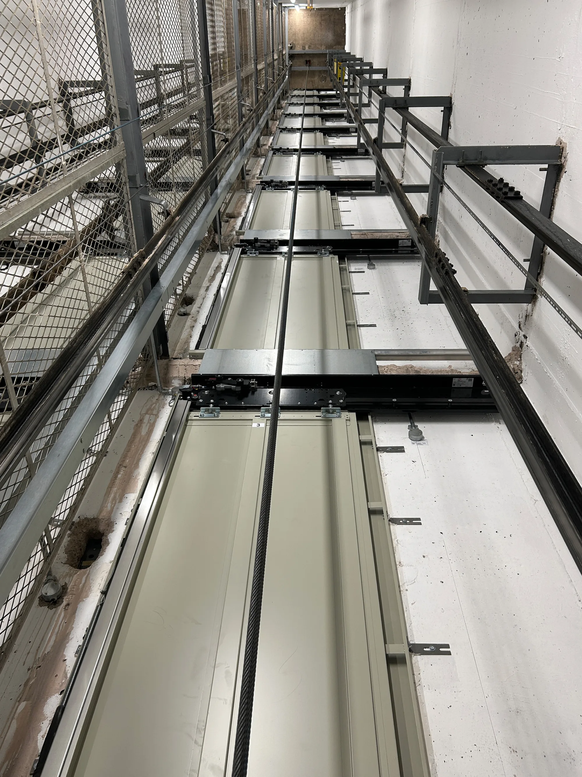 Vertical view inside an elevator shaft showing metal tracks, cables, and structural supports. White walls with visible wear, mesh barrier on the left, and metal framework on the right.