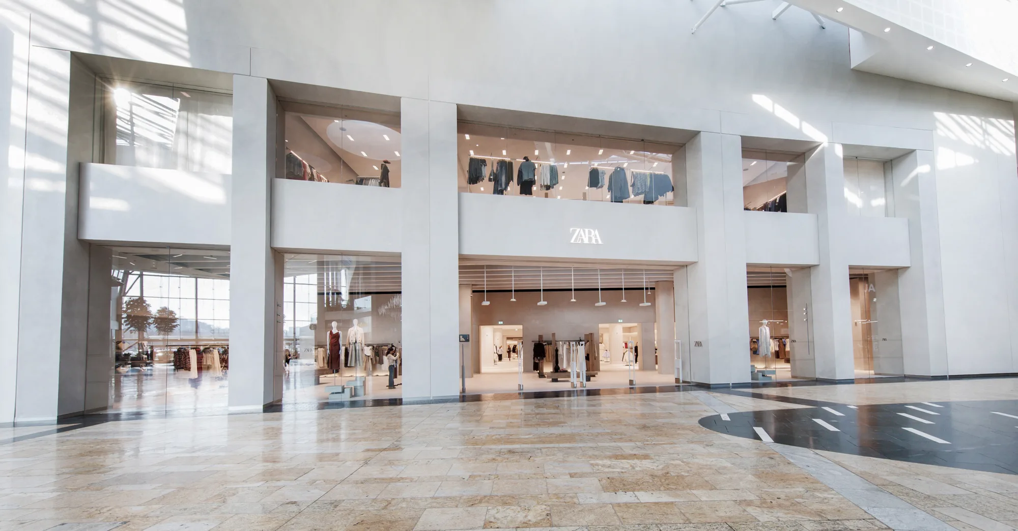 Wide view of a modern shopping mall interior showing a large Zara storefront with three tall glass entrances and upper-level display windows featuring hanging clothing. The floor is polished stone with light and dark sections, and sunlight streams through large windows on the left.