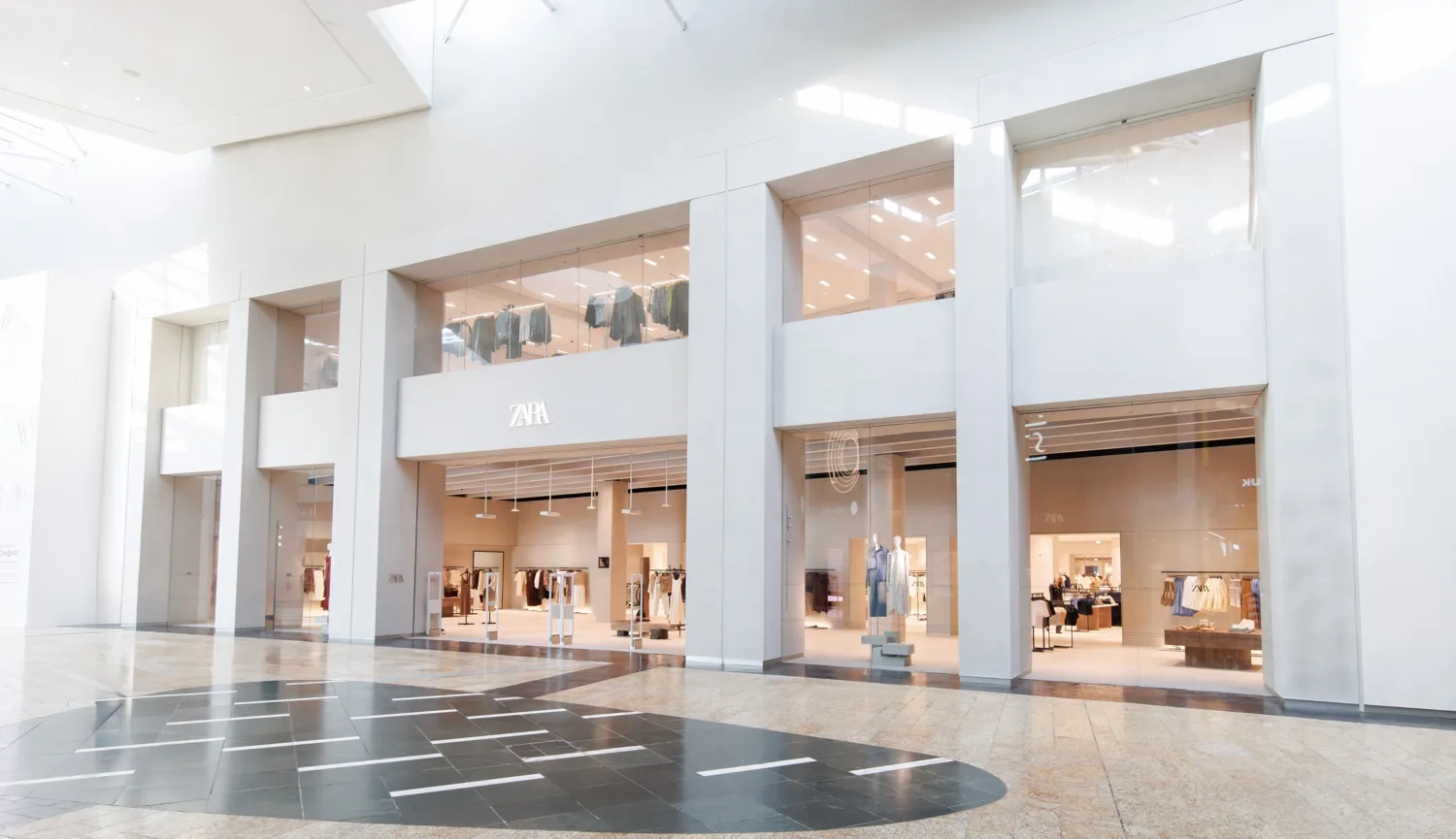 Wide view of a Zara storefront inside a shopping mall, featuring three large glass entrances and upper-level display windows with hanging clothing. The floor has light stone tiles with a dark geometric pattern, and bright natural light enters from the left.