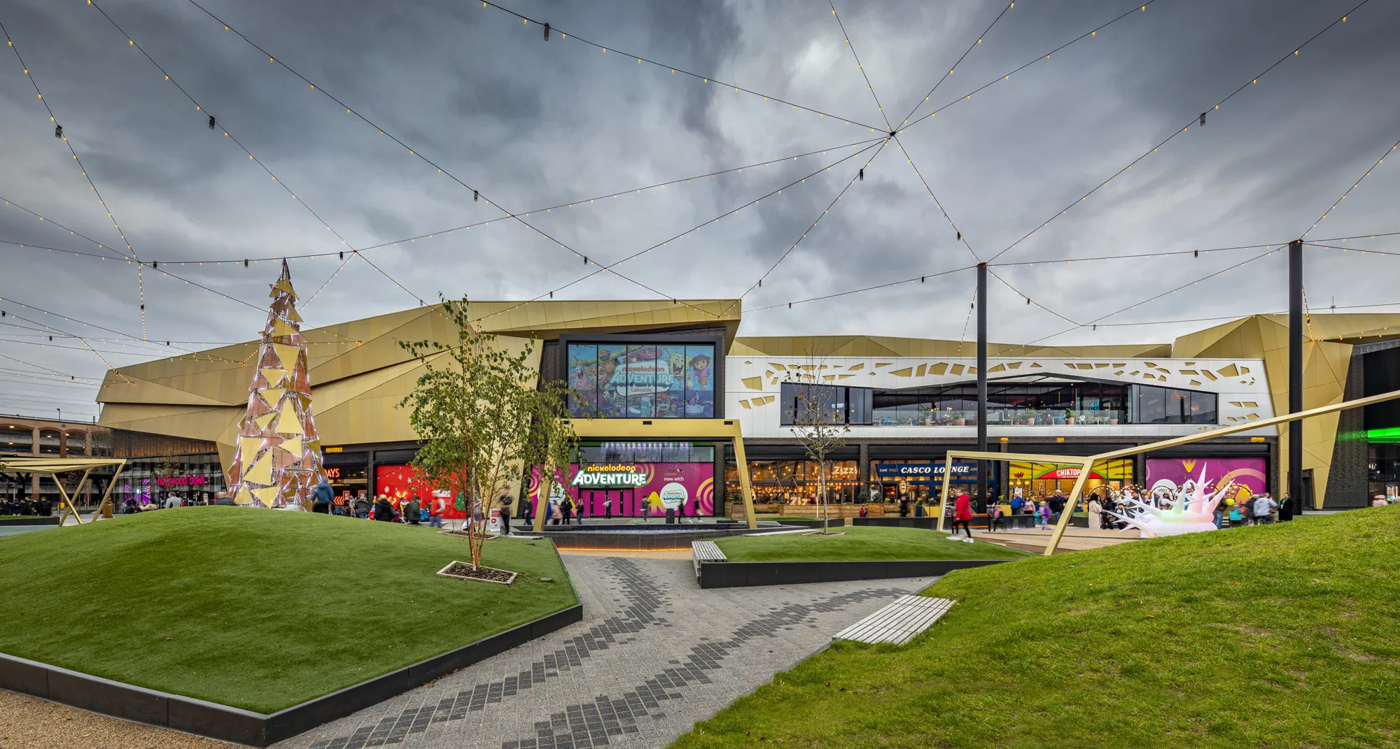 Grass banks with a modern gold christmas tree and suspended fairy lights infront of the gold facade of the intu centre