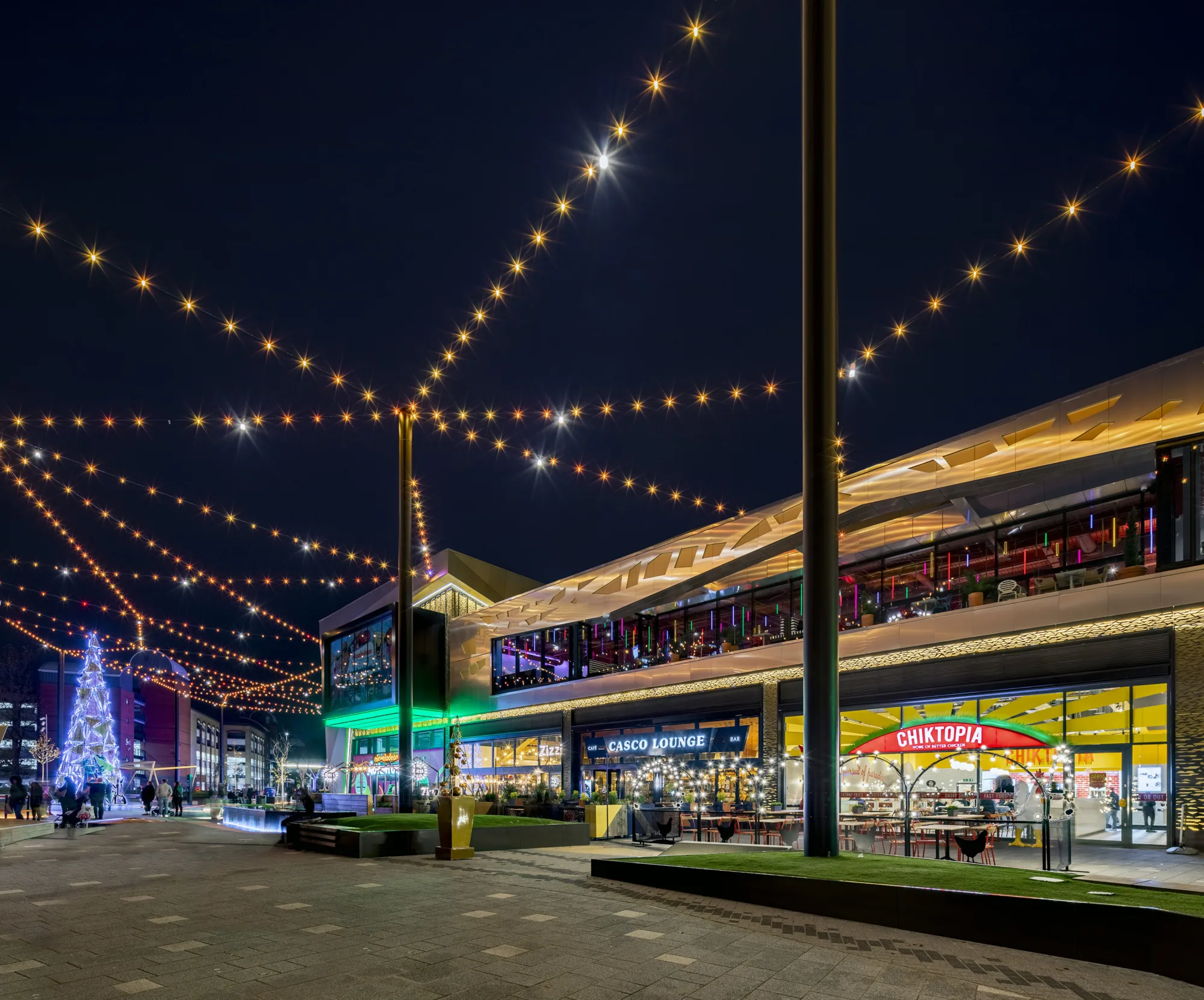 intu at night with restaurant facades and 2nd floor terrace seating. A lit Christmas tree in the background with suspended fairy lights
