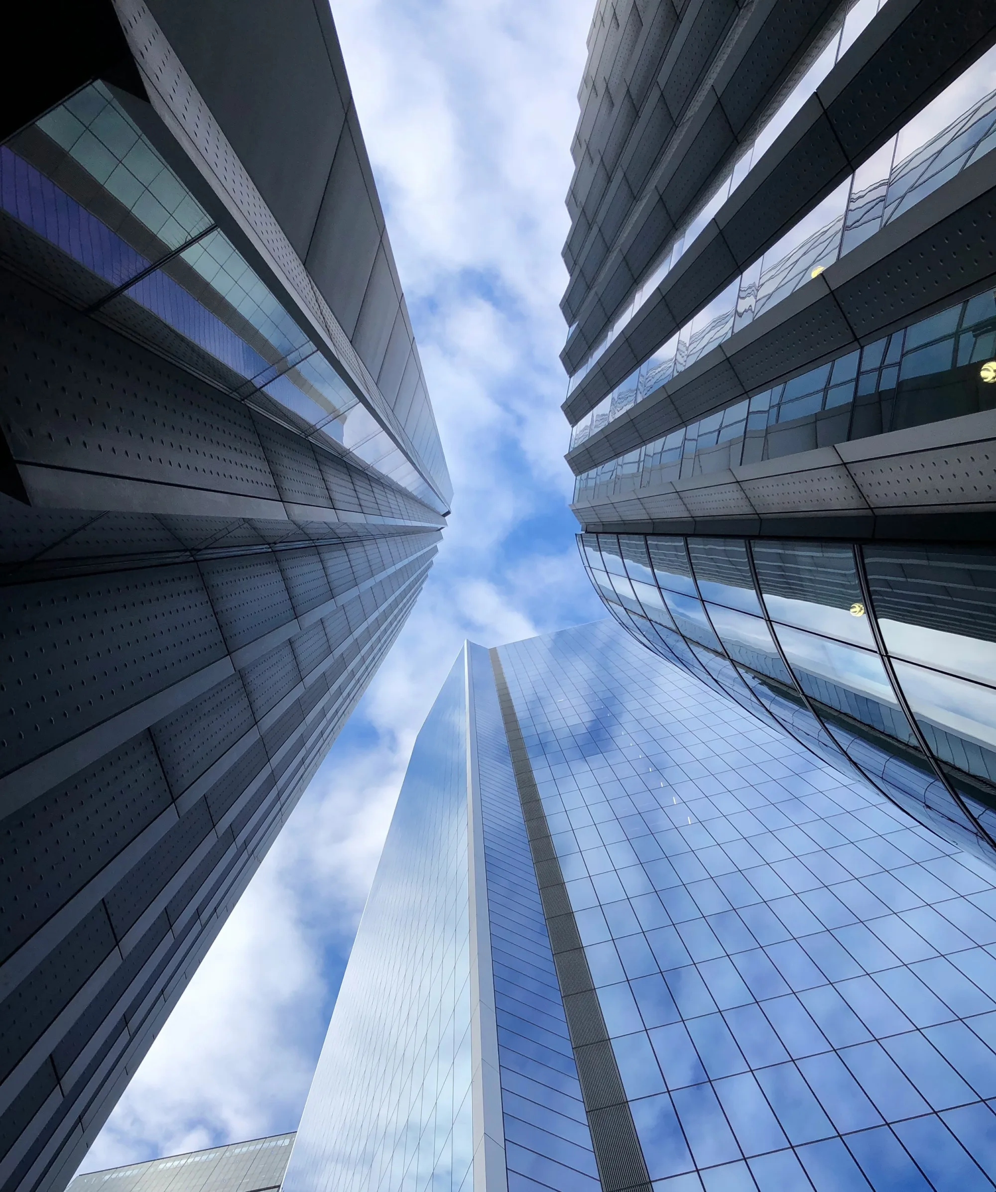 Looking up at glazed highrise buildings into a lightly clouded sky.