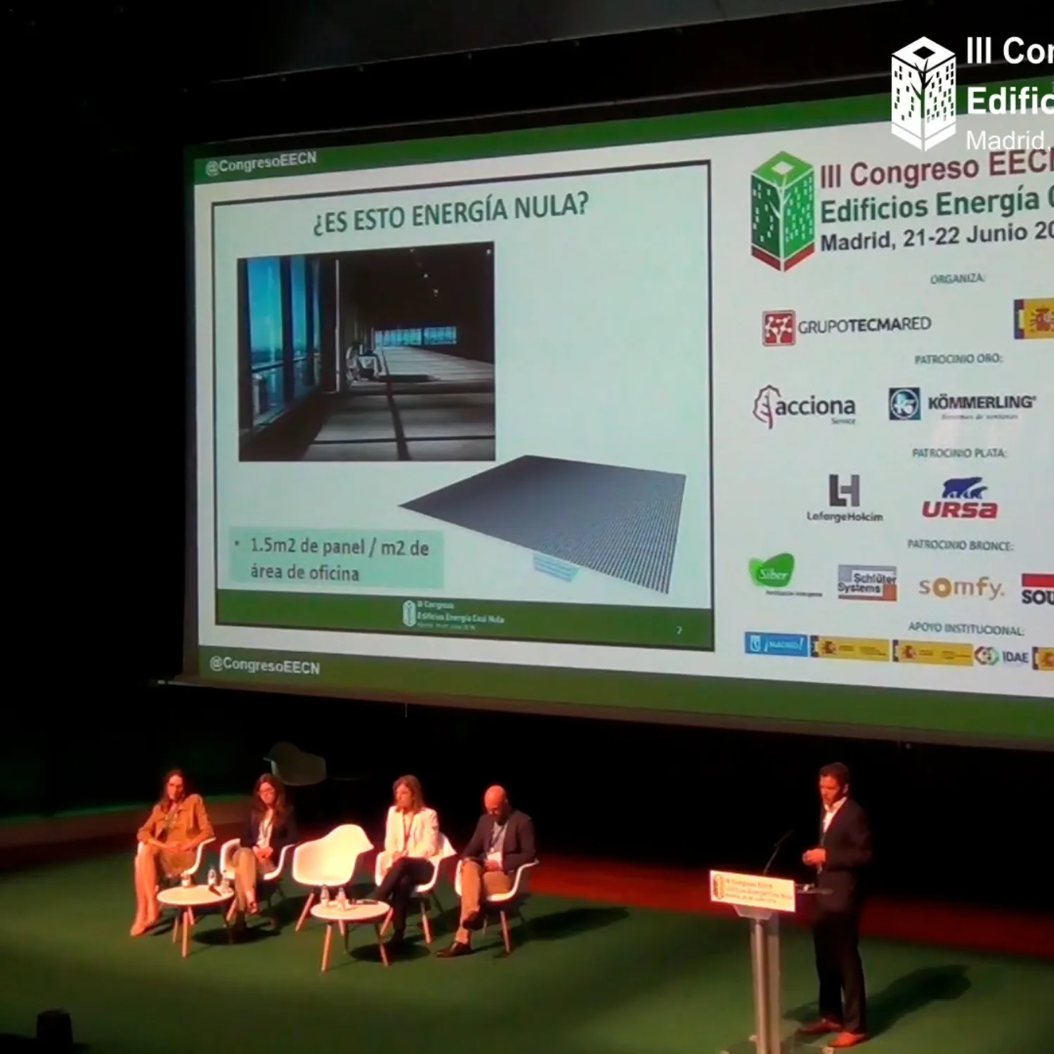Jose on stage with a panel of seated speakers in front of a large projection screen