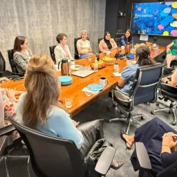 Conference room with a rectangular wooden table surrounded by multiple women seated in office chairs; table has plates, glasses, a coffee pot, and snacks;