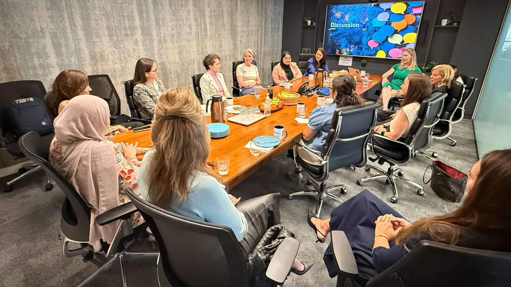 Conference room with a rectangular wooden table surrounded by multiple women seated in office chairs; table has plates, glasses, a coffee pot, and snacks;