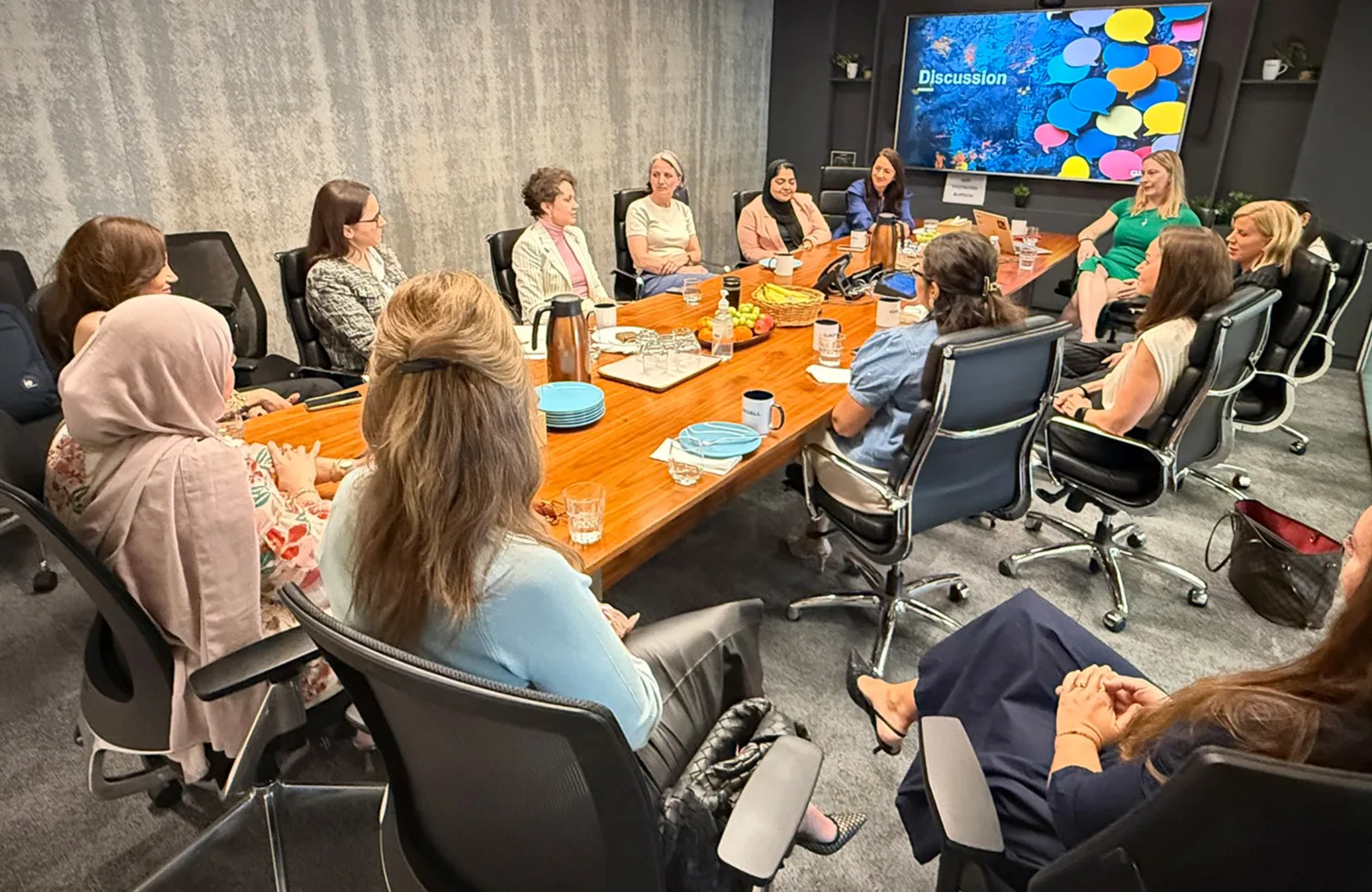 Conference room with a rectangular wooden table surrounded by multiple women seated in office chairs; table has plates, glasses, a coffee pot, and snacks;