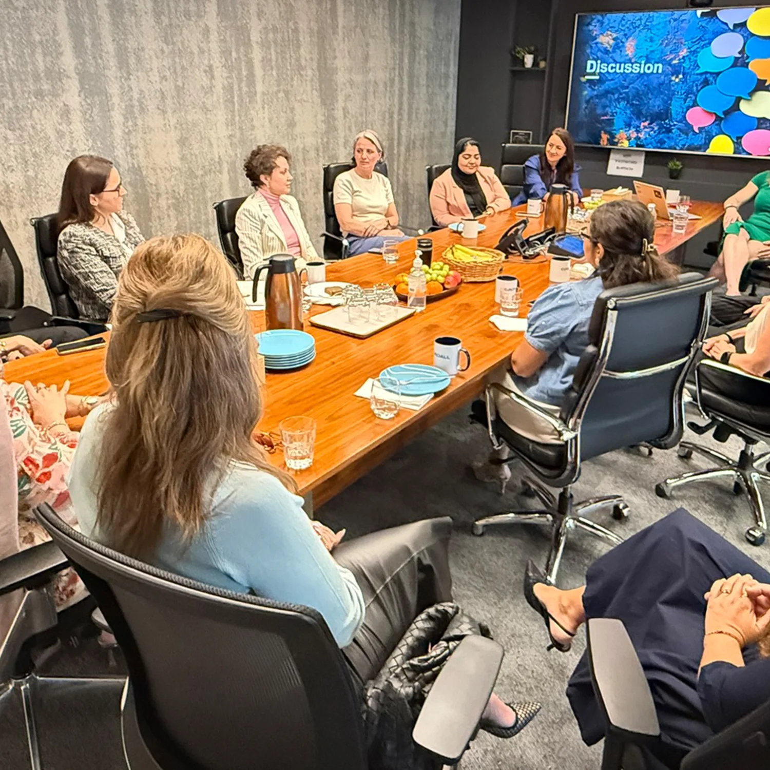 Conference room with a rectangular wooden table surrounded by multiple women seated in office chairs; table has plates, glasses, a coffee pot, and snacks;