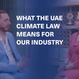 Two people seated across from each other in a professional setting; one in a light blue suit, the other in a red dress. Background shows circular-shelved wall and framed certificates. Text reads: “WHAT THE UAE CLIMATE LAW MEANS FOR OUR INDUSTRY”.
