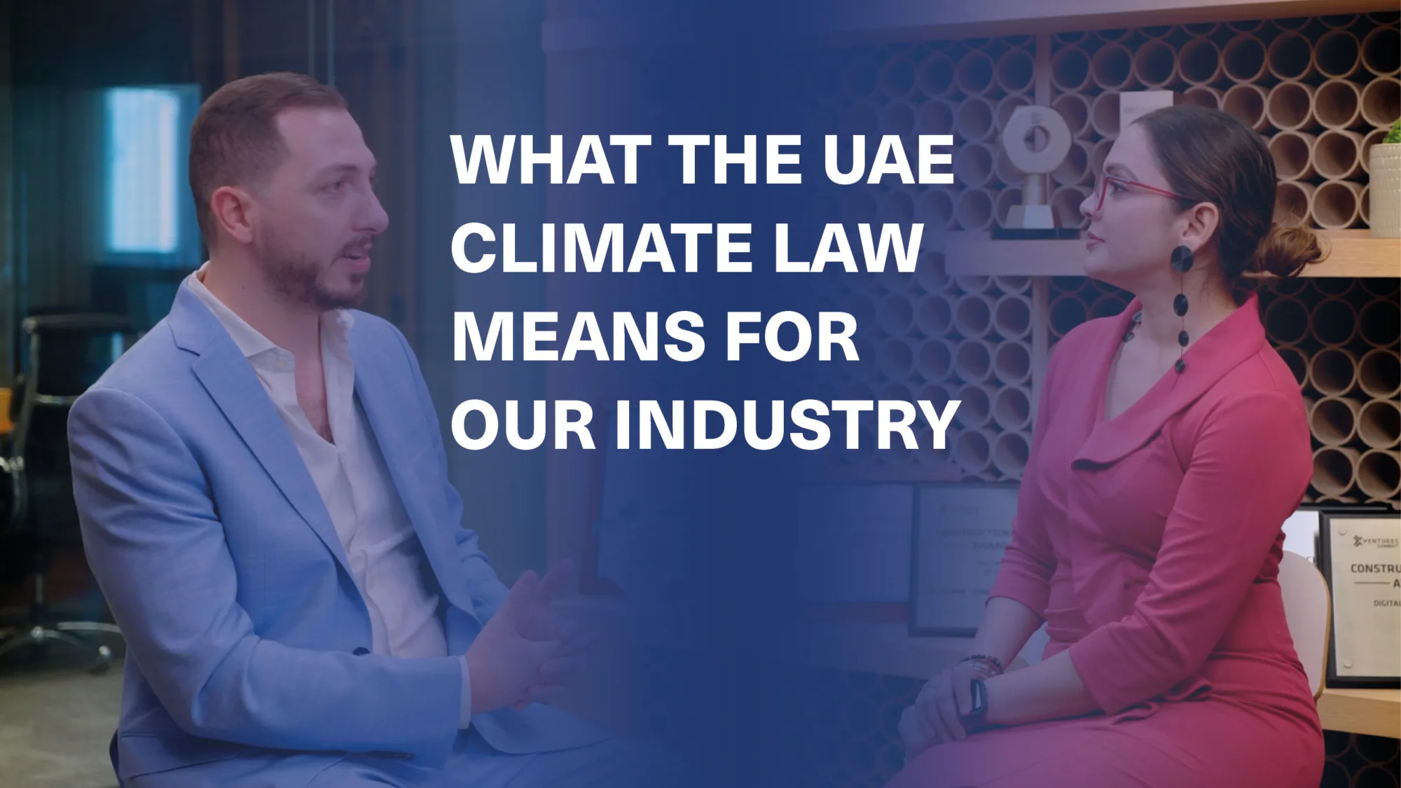 Two people seated across from each other in a professional setting; one in a light blue suit, the other in a red dress. Background shows circular-shelved wall and framed certificates. Text reads: “WHAT THE UAE CLIMATE LAW MEANS FOR OUR INDUSTRY”.