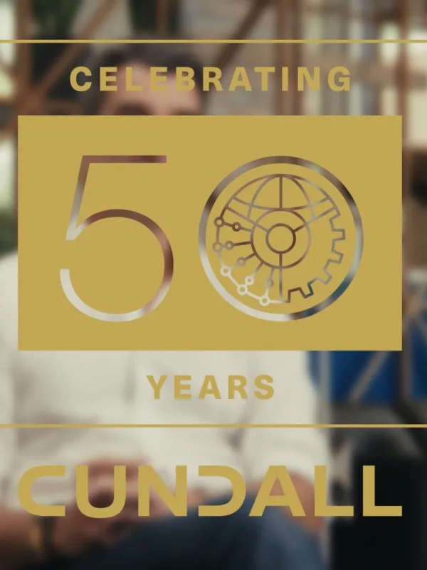 A gold graphic overlay with the text “Celebrating 50 Years Cundall” and a stylised gear and circuit design. The background shows a modern office setting with blue panels, wooden framework, and a black sofa.