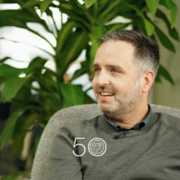 A person wearing a grey sweater sits in an indoor space with large green plants behind them. Part of a window and office interior is visible, and a “50” anniversary-style graphic appears near the bottom of the image.