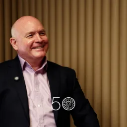 A person in a dark suit jacket and pale pink shirt sits against a backdrop of vertical tan acoustic panels. A round lapel pin is visible on the jacket, and a “50” anniversary-style graphic appears near the bottom of the image.