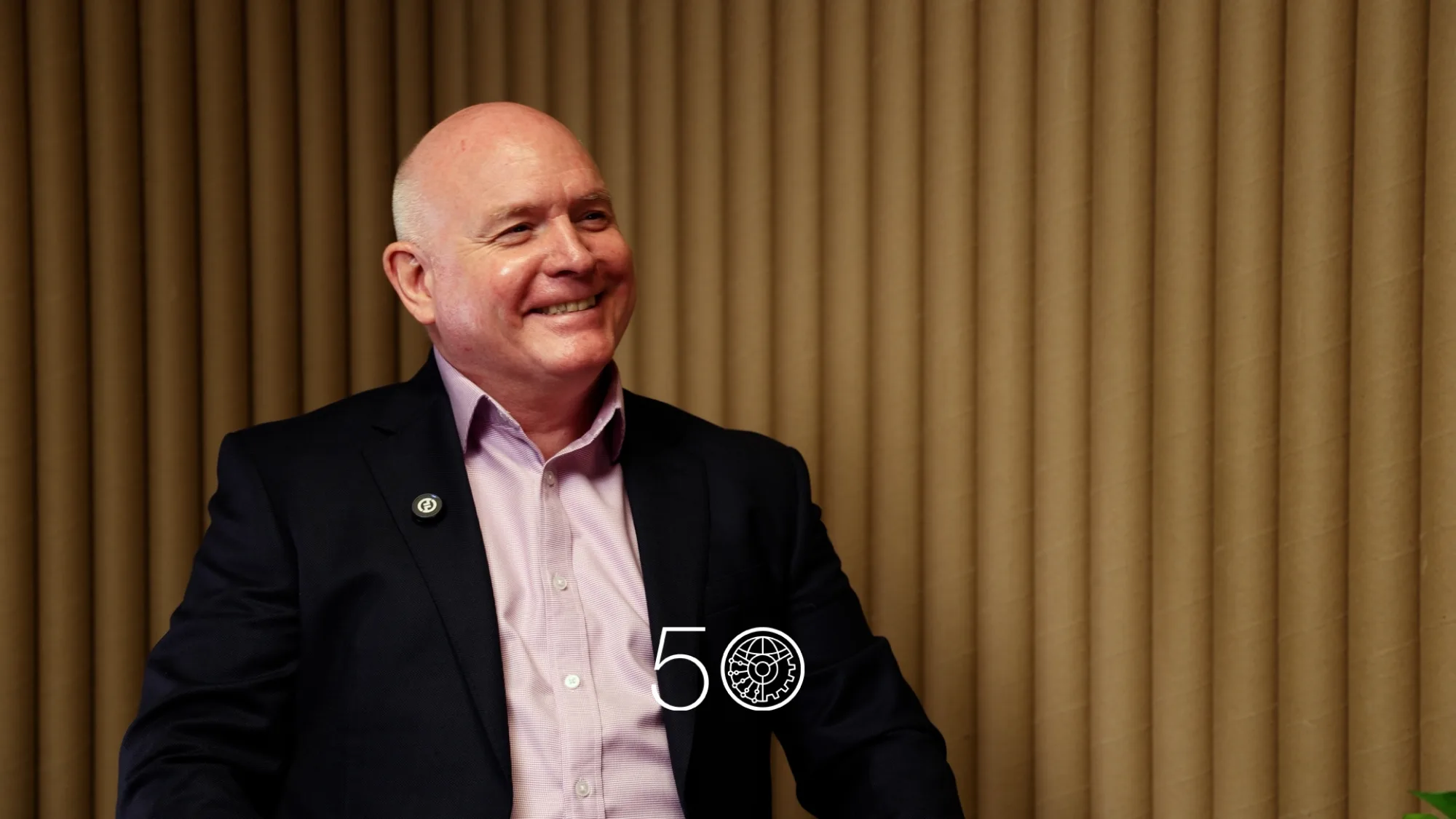 A person in a dark suit jacket and pale pink shirt sits against a backdrop of vertical tan acoustic panels. A round lapel pin is visible on the jacket, and a “50” anniversary-style graphic appears near the bottom of the image.
