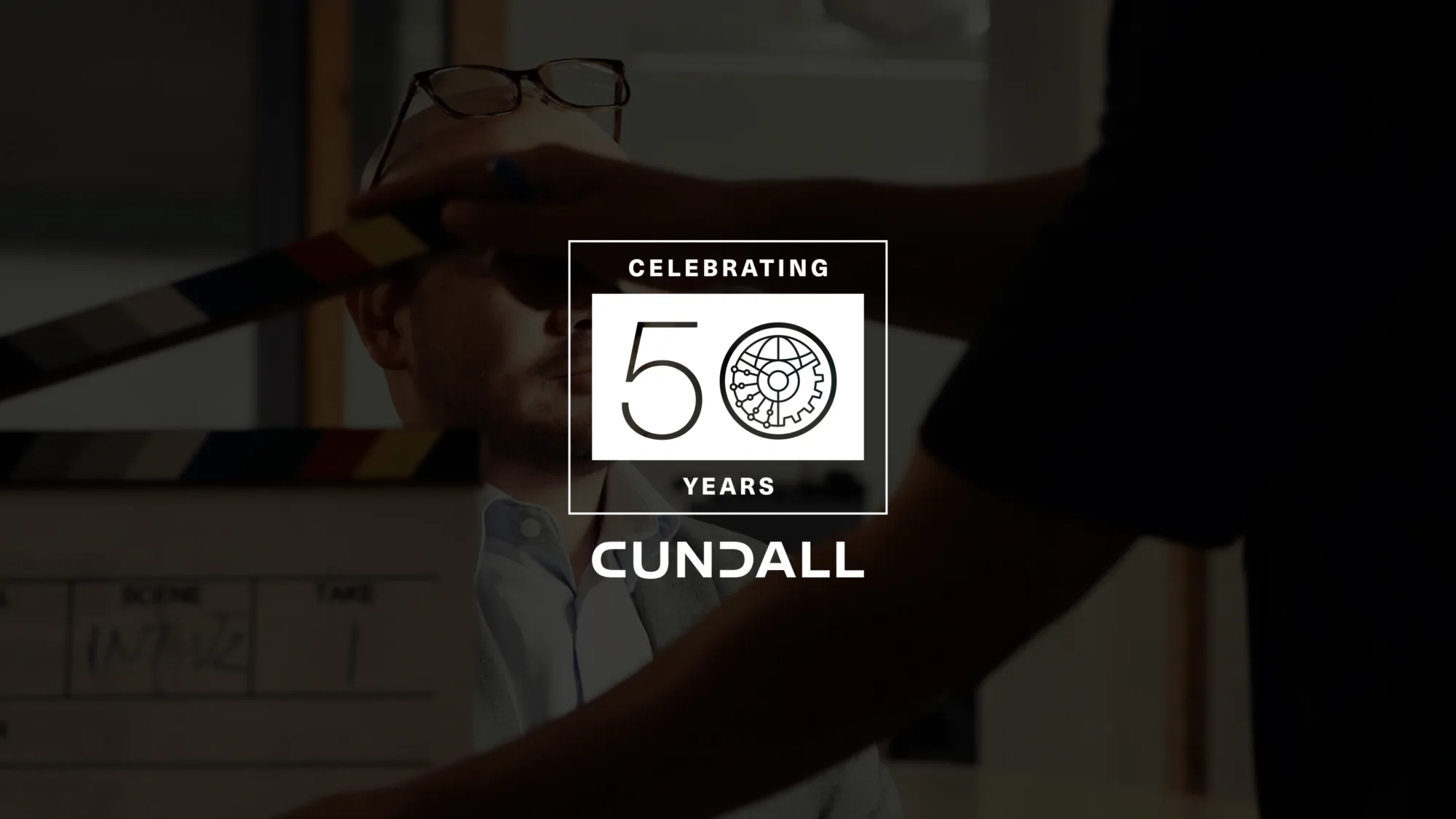 A person seated indoors with glasses resting on their head, while another person holds a clapperboard in front of them. Overlaid text reads “Celebrating 50 Years Cundall” with a gear icon in the design.