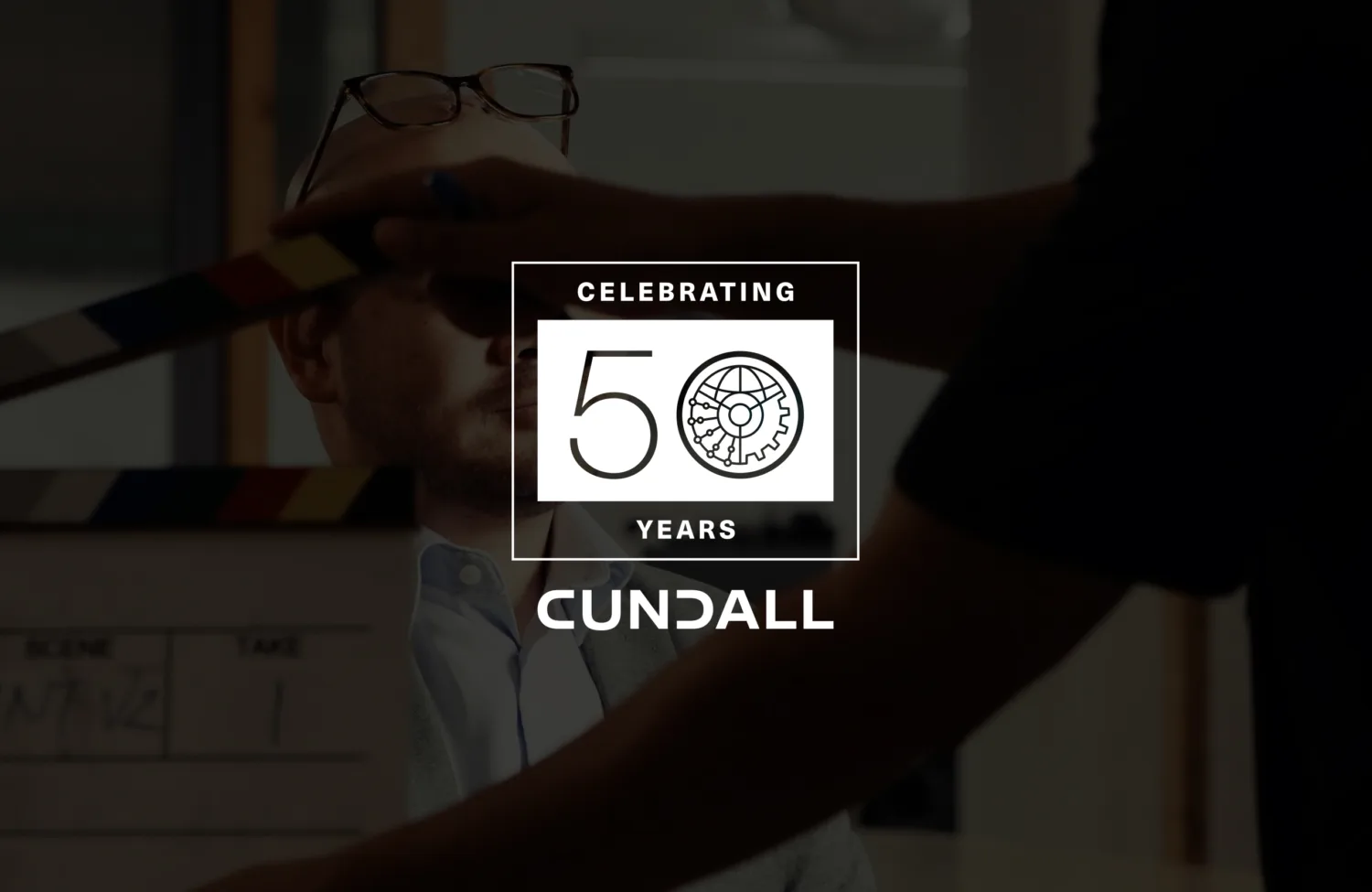A person seated indoors with glasses resting on their head, while another person holds a clapperboard in front of them. Overlaid text reads “Celebrating 50 Years Cundall” with a gear icon in the design.