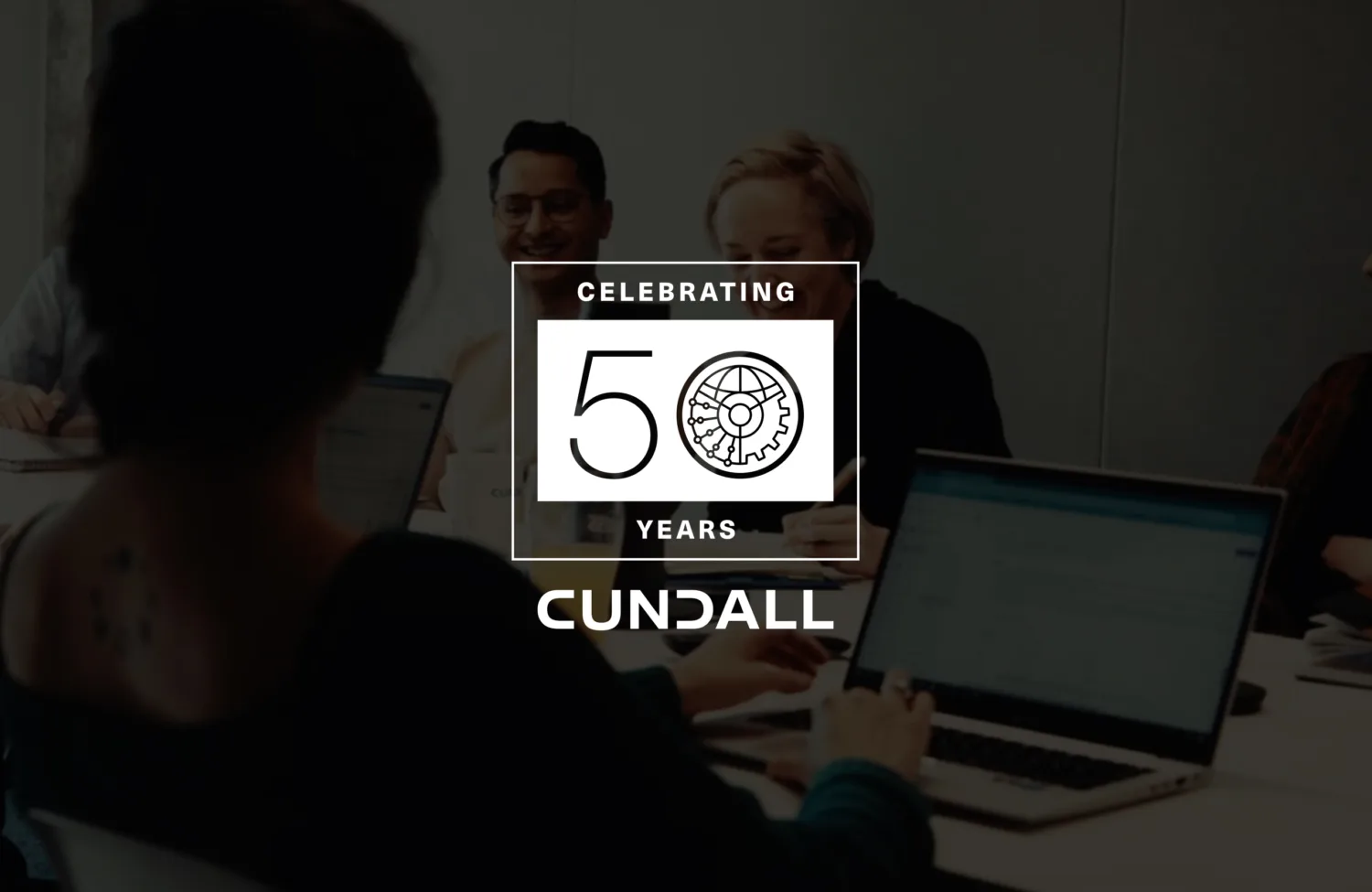 A group of people seated around a meeting table with laptops and notebooks in a modern office setting. Overlaid graphic reads “Celebrating 50 Years Cundall” with a gear and circuit design.