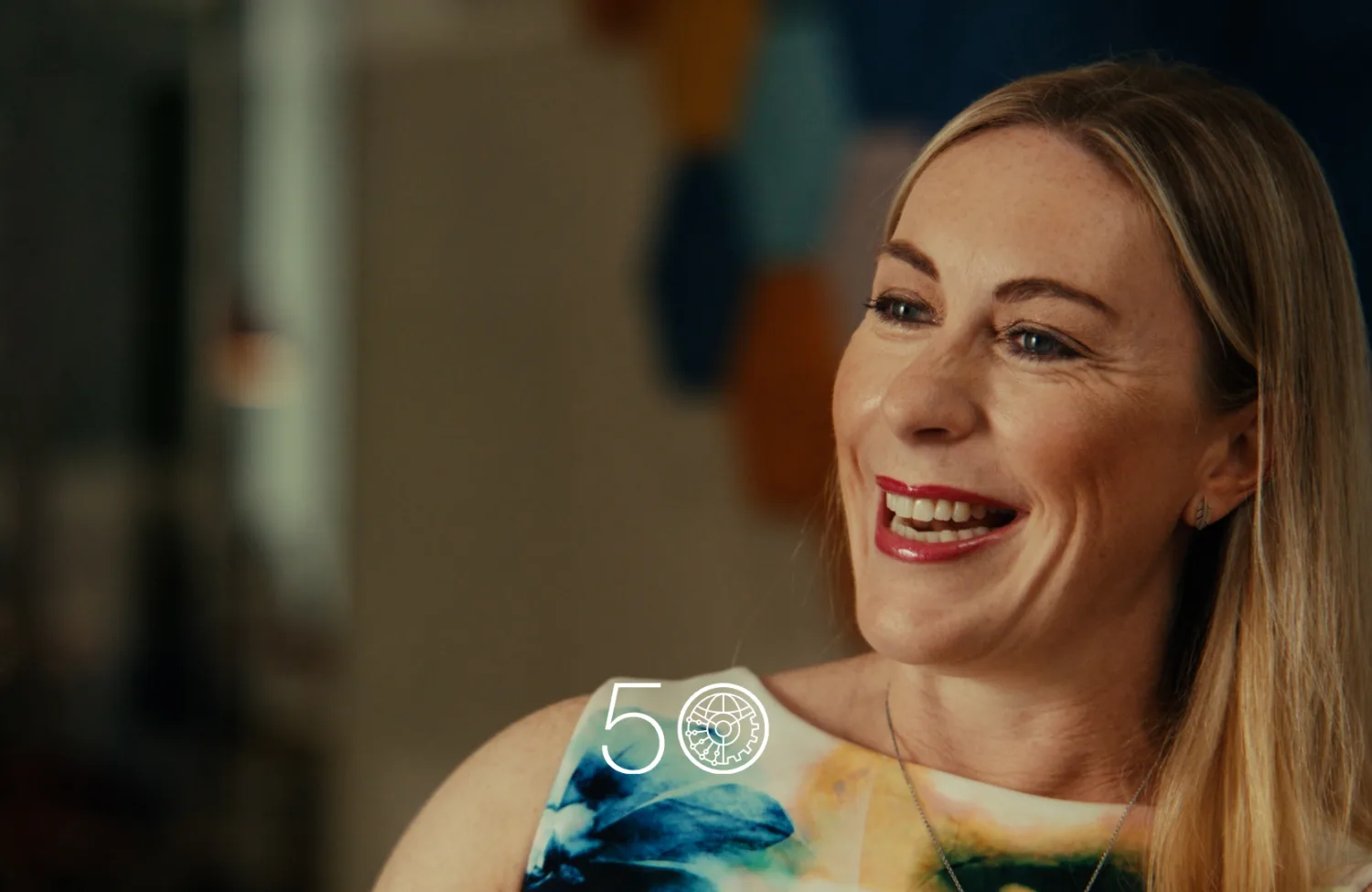 A person with long blond hair sits indoors wearing a sleeveless top with a blue, green, and yellow abstract pattern. The background is softly blurred, and a “50” anniversary-style graphic appears near the bottom of the image.