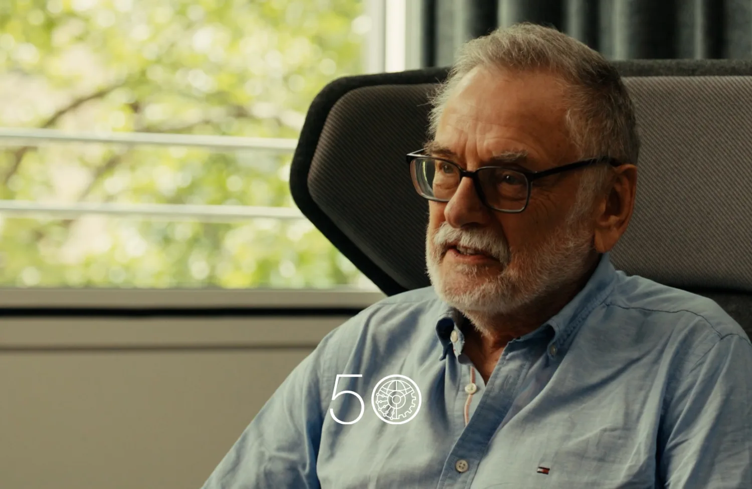 A person in a light blue button-up shirt sits in a modern chair near a large window with green trees visible outside. Dark curtains hang to the right, and a “50” anniversary-style graphic appears at the bottom of the image.
