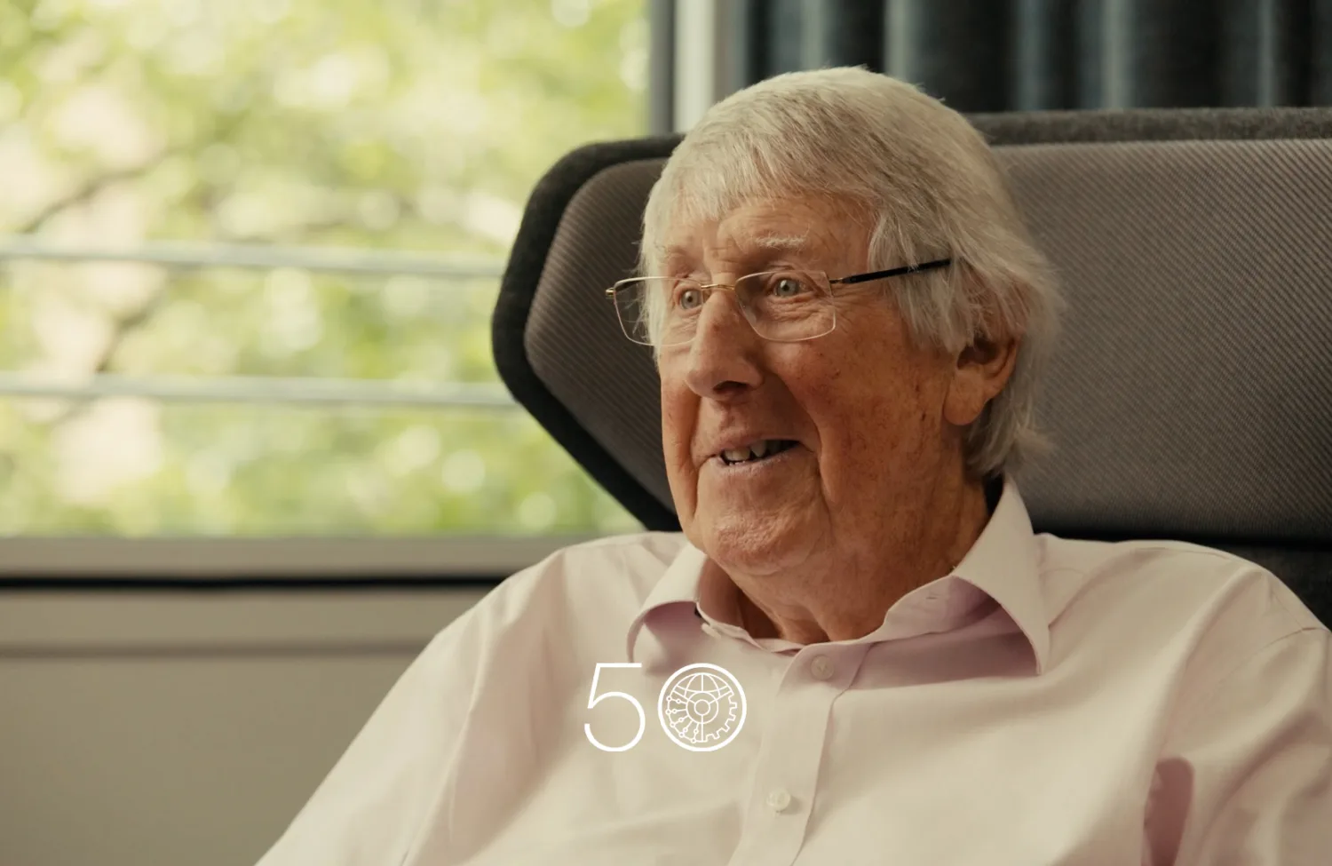 A person wearing a light pink button-up shirt sits in a cushioned chair near a window with green trees visible outside. Dark curtains frame part of the background, and a “50” anniversary-style graphic appears at the bottom of the image.