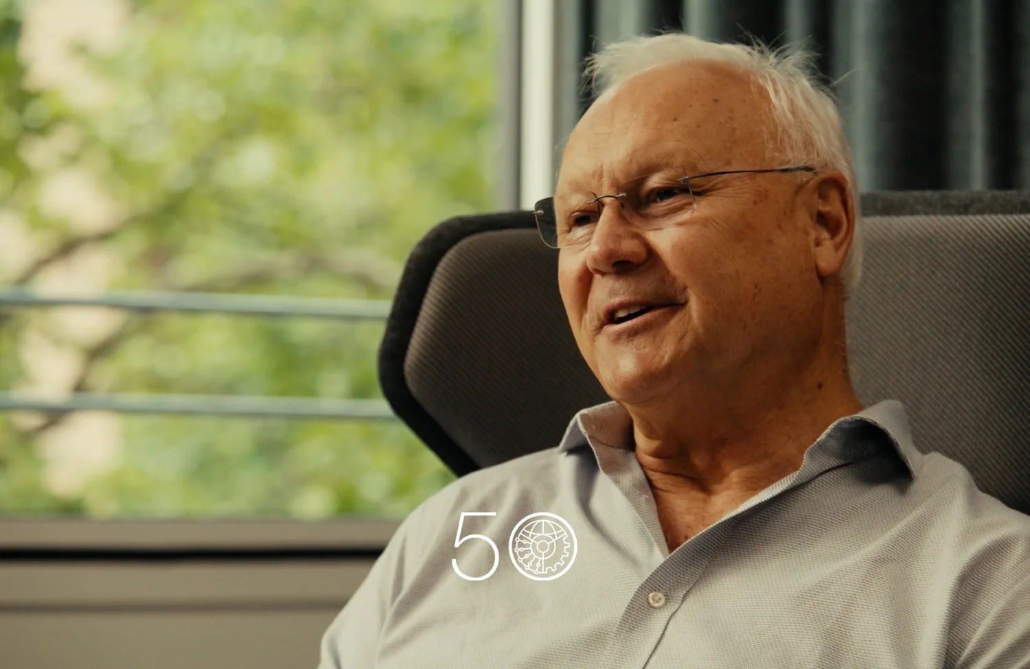 A person in a light grey button-up shirt sits in a cushioned chair near a large window with green trees visible outside. Dark curtains frame part of the background, and a “50” anniversary-style graphic appears at the bottom of the image.