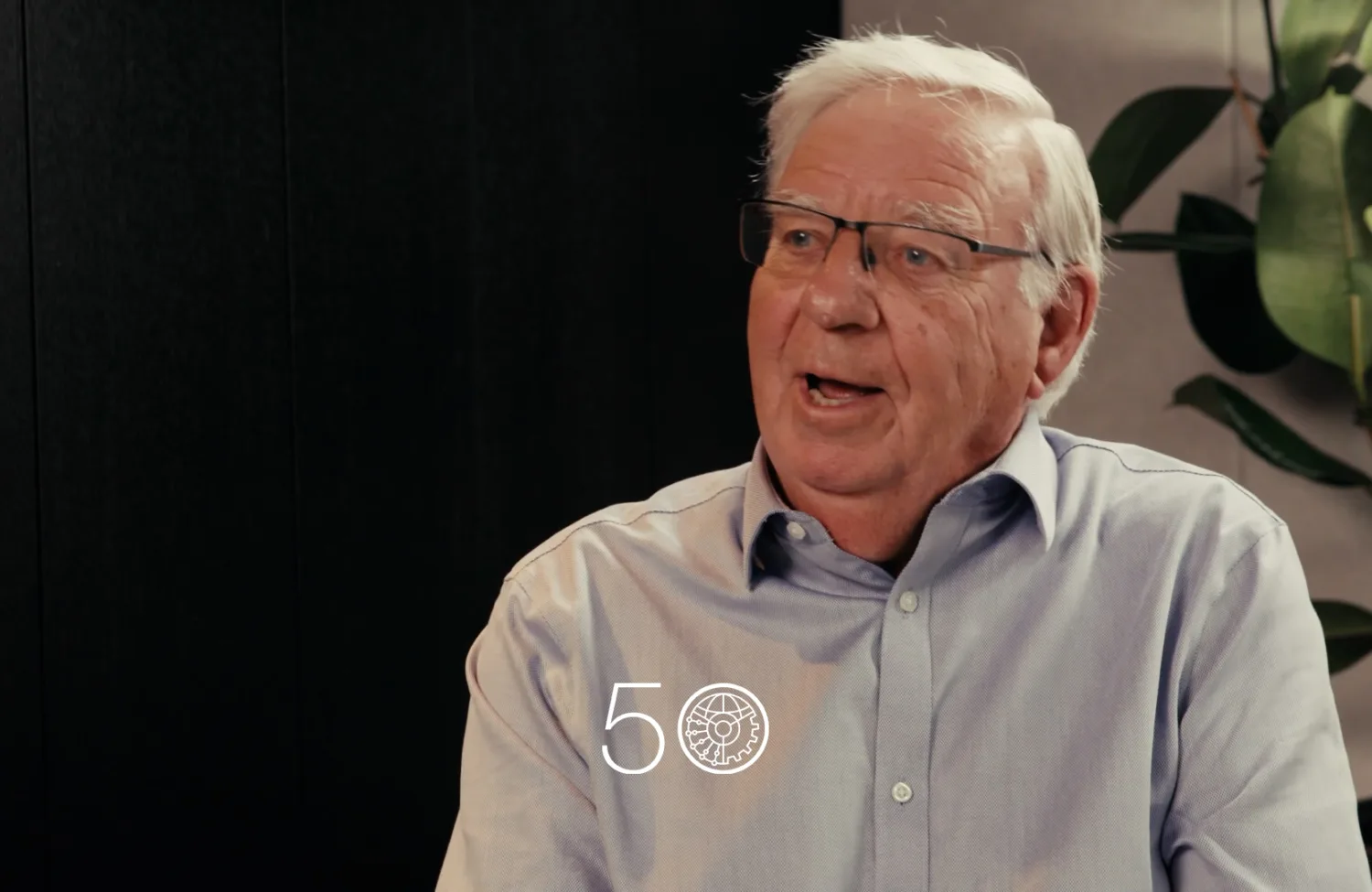 A person wearing a light blue button-up shirt sits indoors against a dark wall. A tall plant with broad green leaves is visible to the right, and a “50” anniversary-style graphic appears near the bottom of the image.