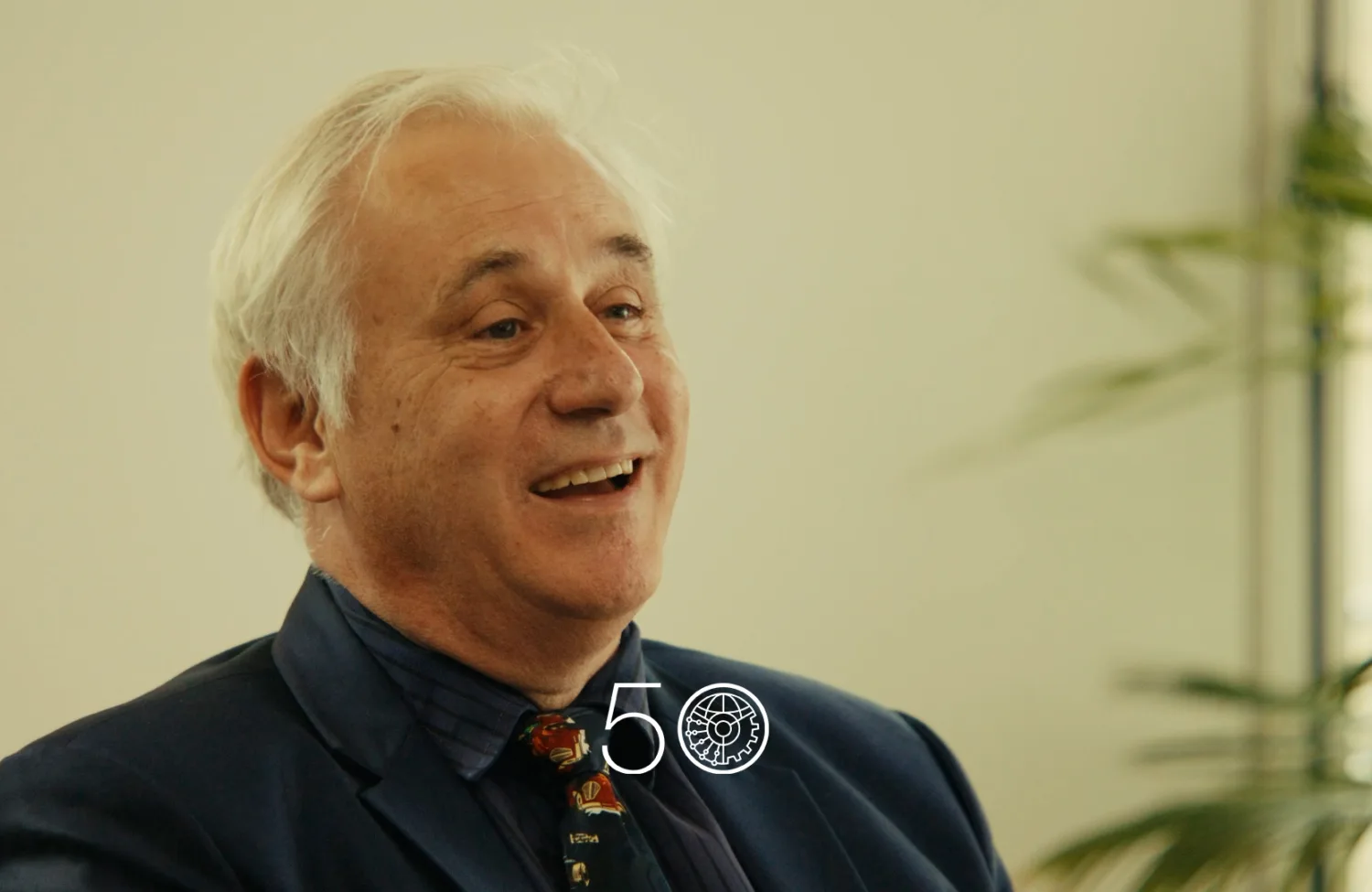 A person wearing a dark suit jacket, navy shirt, and patterned tie sits indoors against a light wall. Green plants frame the right side of the scene, and a “50” anniversary-style graphic appears at the bottom of the image.