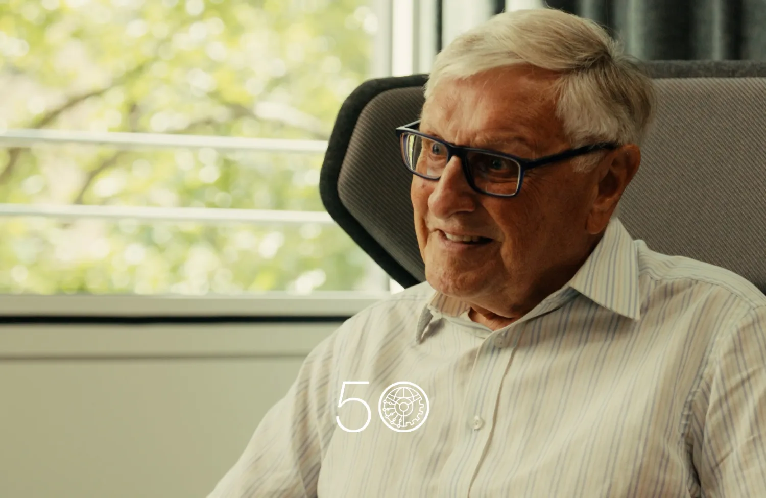 A person wearing a light striped button-up shirt sits in a cushioned chair beside a large window with green trees visible outside. Dark curtains frame part of the background, and a “50” anniversary-style graphic appears at the bottom of the image.