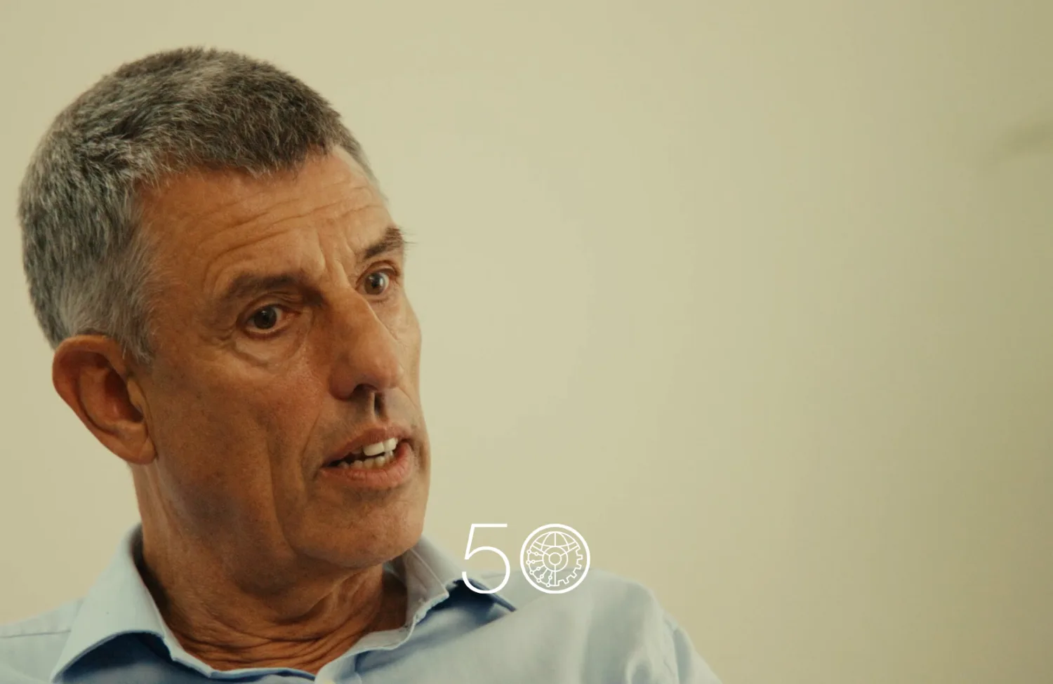 A person in a light blue collared shirt sits indoors against a plain light-colored wall. Part of a green plant is visible on the right, and a “50” anniversary-style graphic appears near the bottom of the image.