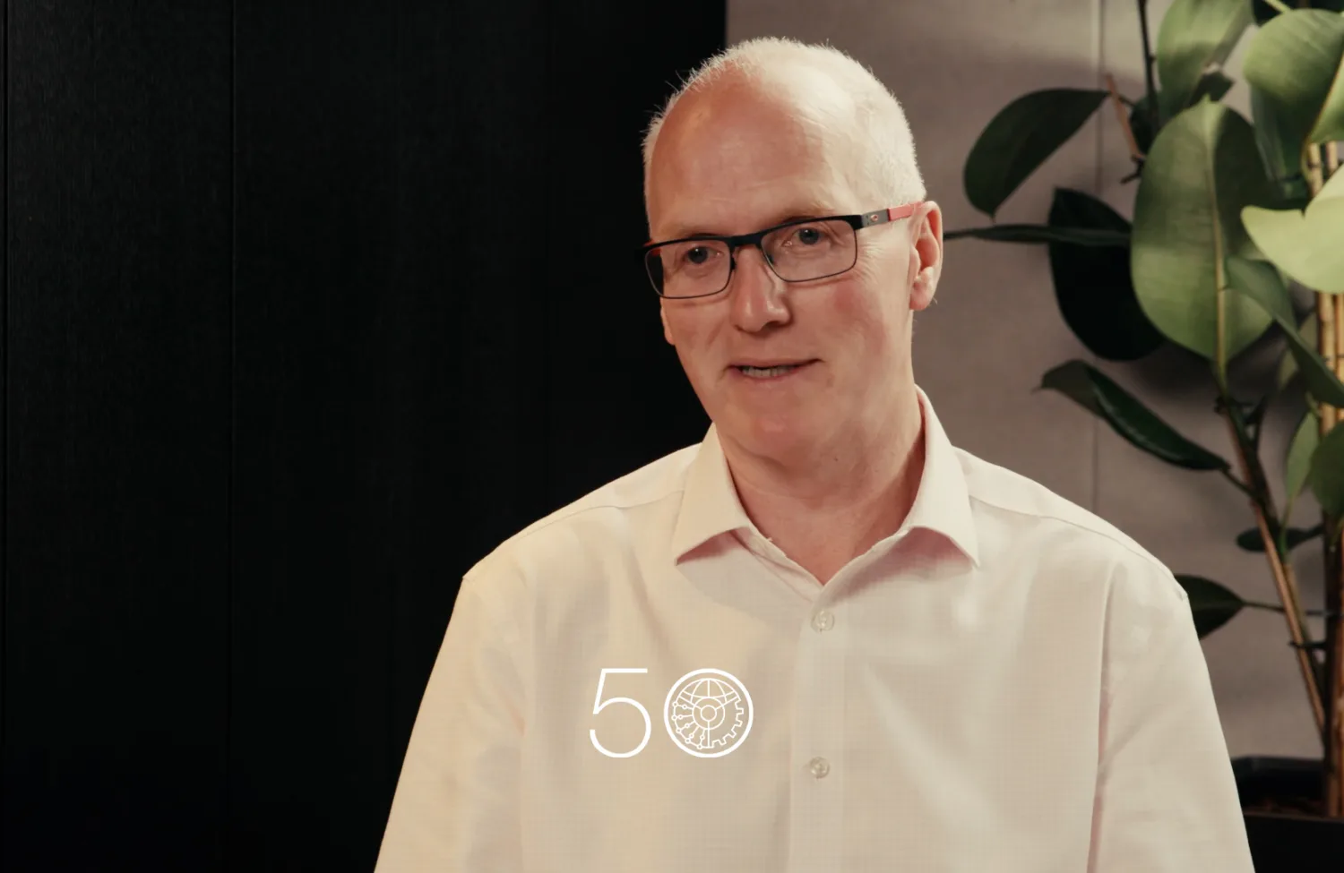 A person wearing a light pink button-up shirt sits indoors against a dark wall, with a tall green plant on the right. A “50” anniversary-style graphic is displayed near the bottom of the image.