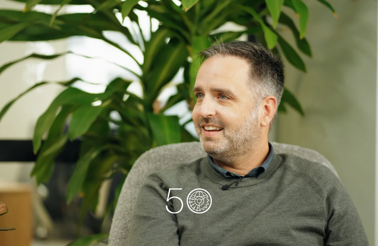 A person wearing a grey sweater sits in an indoor space with large green plants behind them. Part of a window and office interior is visible, and a “50” anniversary-style graphic appears near the bottom of the image.