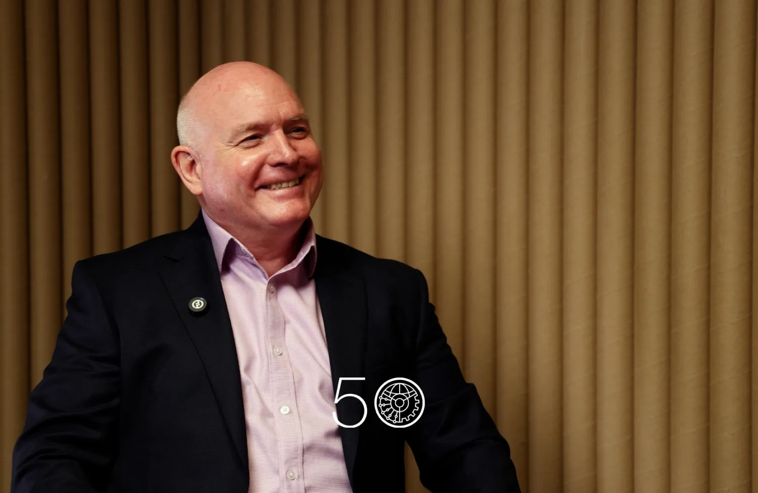A person in a dark suit jacket and pale pink shirt sits against a backdrop of vertical tan acoustic panels. A round lapel pin is visible on the jacket, and a “50” anniversary-style graphic appears near the bottom of the image.