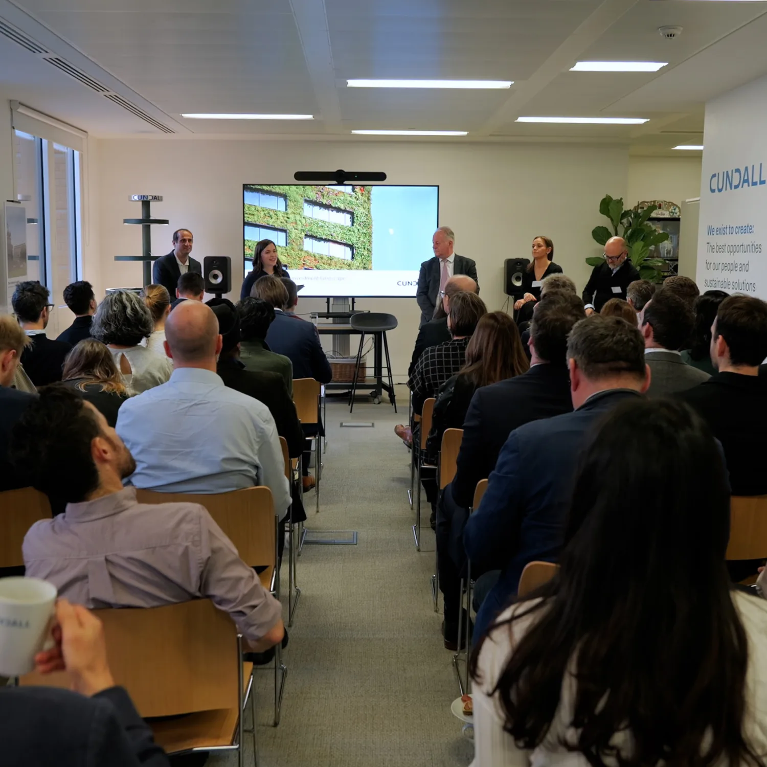 Backs of heads of the seated audience with the panel stood in front of the screen presentation