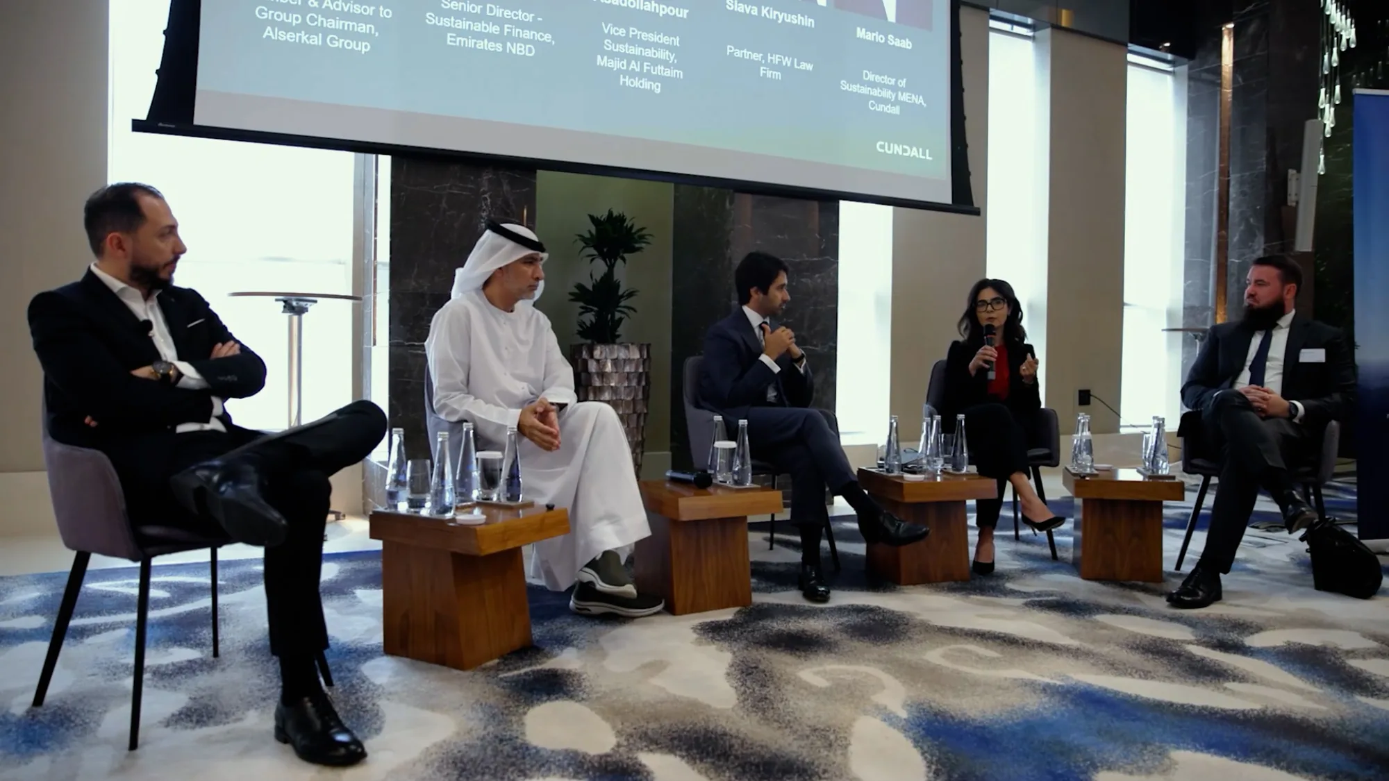 Five individuals seated on chairs in a conference room for a panel discussion; large screen behind them displays names and titles of speakers; microphones and water bottles on small tables in front.