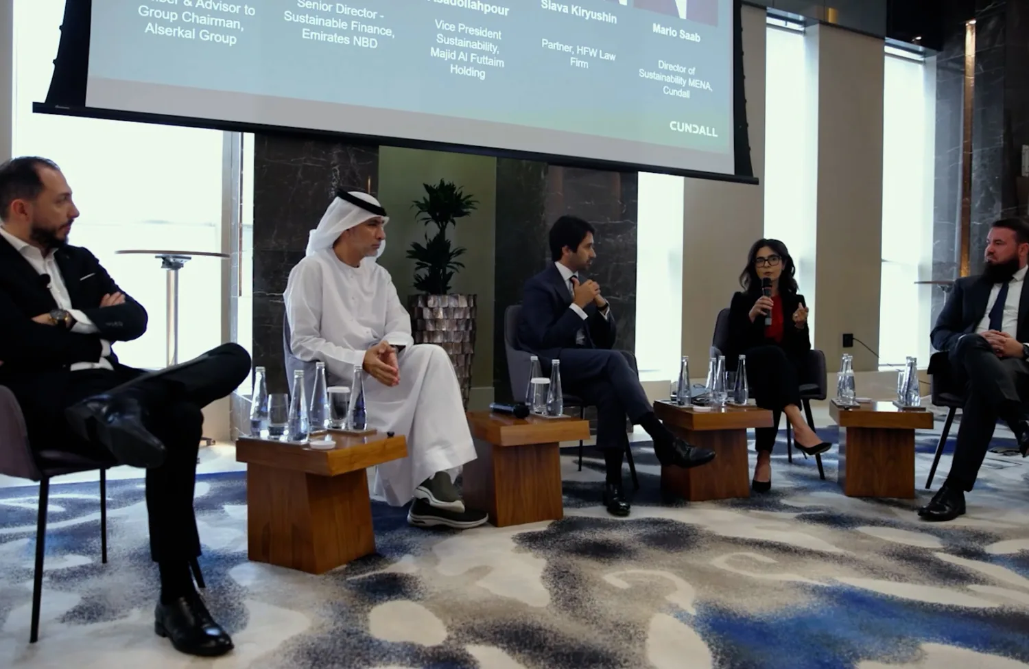 Five individuals seated on chairs in a conference room for a panel discussion; large screen behind them displays names and titles of speakers; microphones and water bottles on small tables in front.