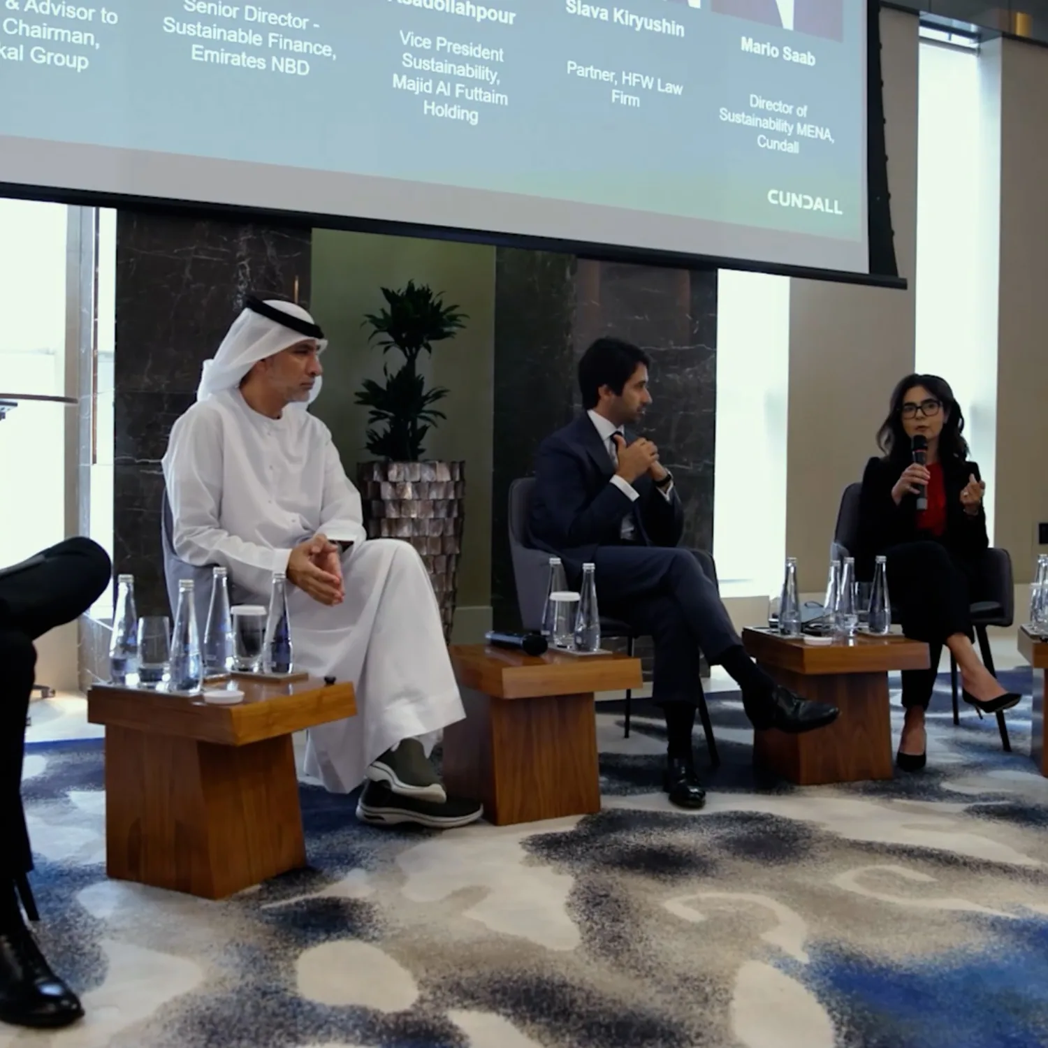 Five individuals seated on chairs in a conference room for a panel discussion; large screen behind them displays names and titles of speakers; microphones and water bottles on small tables in front.