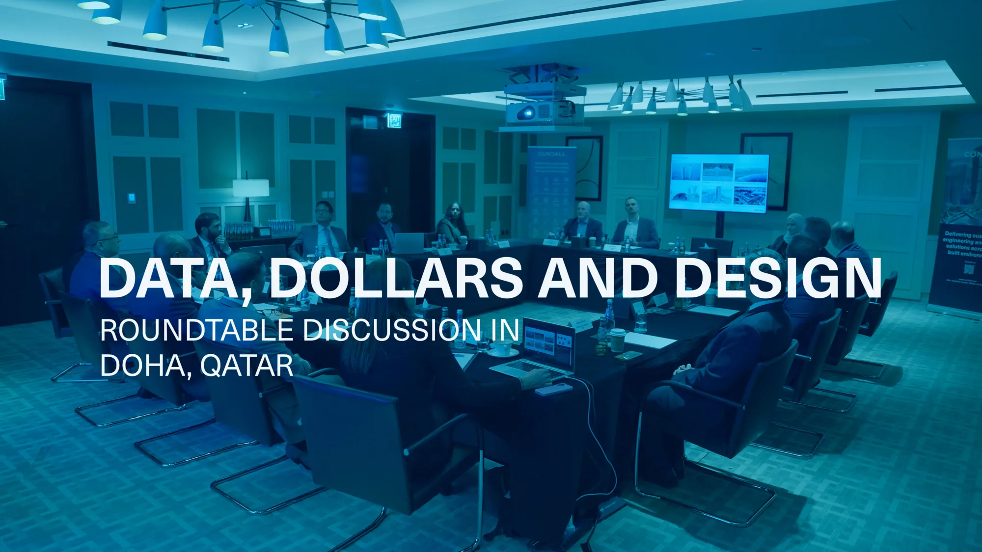 Conference room with a roundtable discussion in progress; several individuals seated around a rectangular table with laptops and documents; large screen at the front displays presentation slides; visible text overlay reads “DATA, DOLLARS AND DESIGN – ROUNDTABLE DISCUSSION IN DOHA, QATAR.