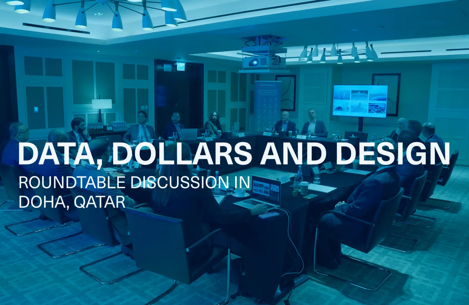 Conference room with a roundtable discussion in progress; several individuals seated around a rectangular table with laptops and documents; large screen at the front displays presentation slides; visible text overlay reads “DATA, DOLLARS AND DESIGN – ROUNDTABLE DISCUSSION IN DOHA, QATAR.