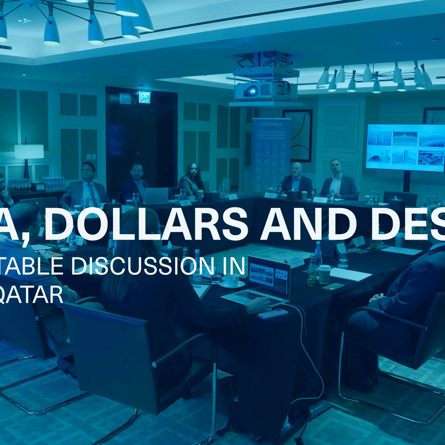 Conference room with a roundtable discussion in progress; several individuals seated around a rectangular table with laptops and documents; large screen at the front displays presentation slides; visible text overlay reads “DATA, DOLLARS AND DESIGN – ROUNDTABLE DISCUSSION IN DOHA, QATAR.