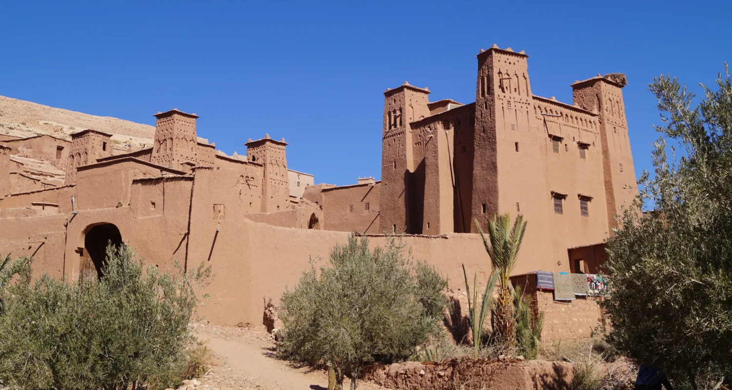 Red sand and brick building in the middle east with rugs drying on the wall behind green palm trees.